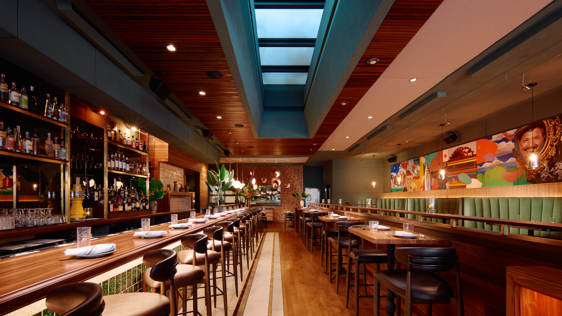 Interior of a stylish restaurant with a high ceiling featuring skylights, a long wooden bar with barstools, and a mural with vibrant artwork on the wall.