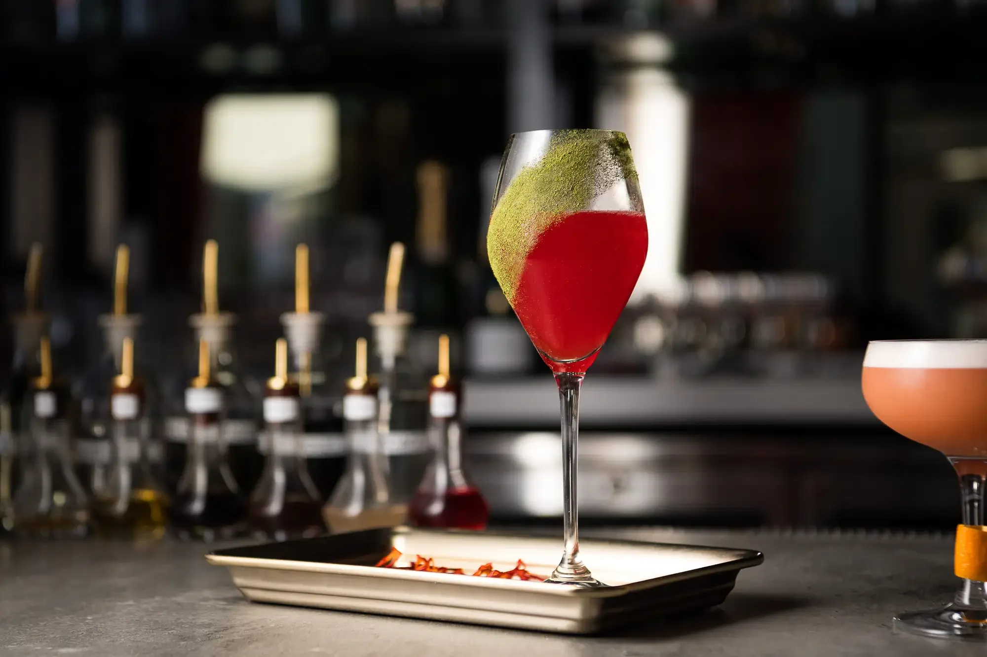 A cocktail in a tall, curvy glass with red liquid and green powder garnish