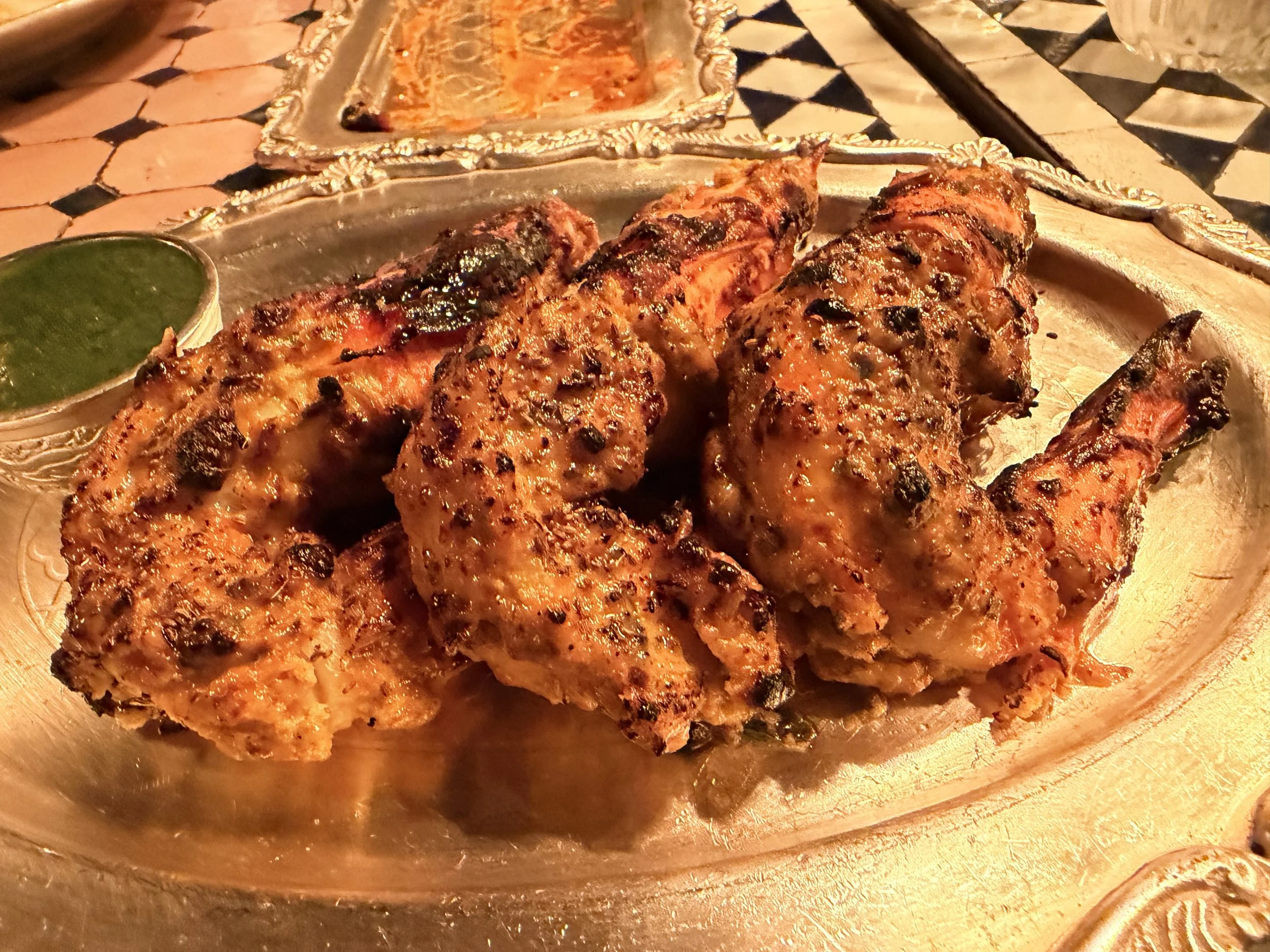 Close-up of grilled jumbo prawns with a spicy, seasoned glaze on a silver platter, with a small cup of green sauce on the side.
