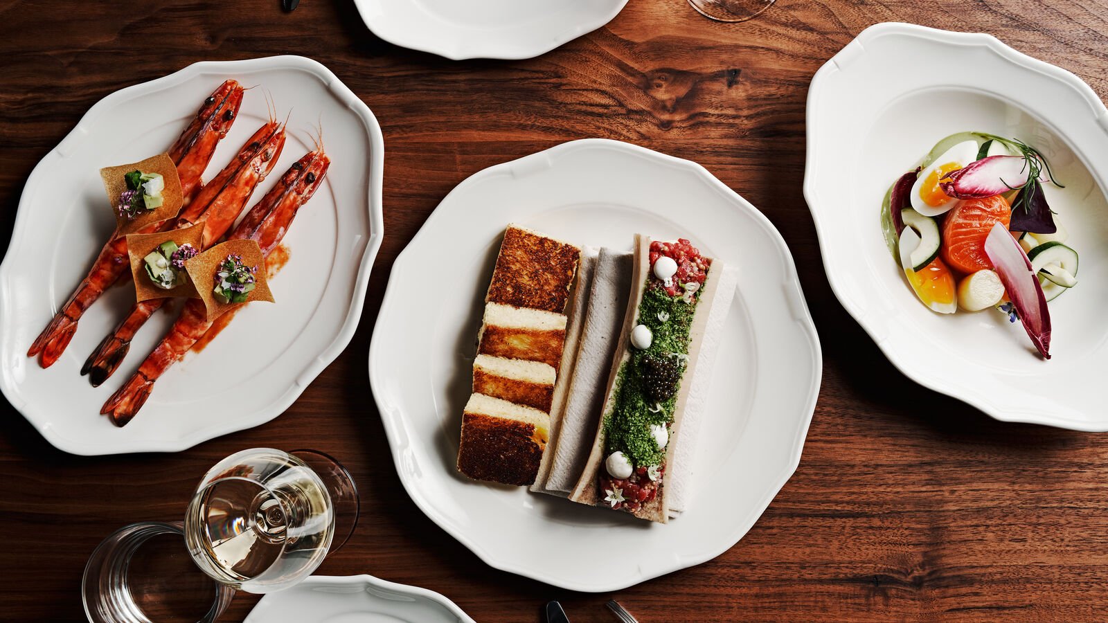 Elegant white plates on a wooden table containing a shrimp appetizer, colorful layered sandwiches, and a fresh salad with smoked salmon, vegetables, and boiled eggs.