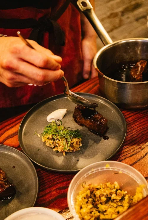 Chef garnishing a plated dish with sauce in a restaurant setting.