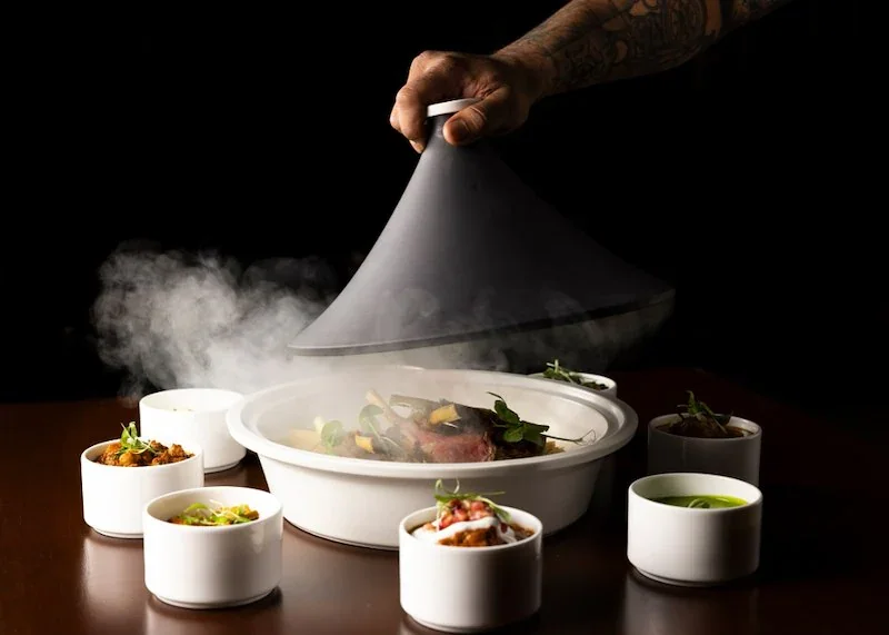Chef lifting a domed cover off a steaming dish surrounded by small white bowls of food.