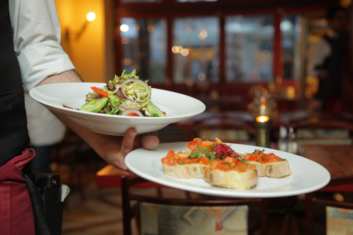Person holding two white plates with salads and bruschetta inside a restaurant.