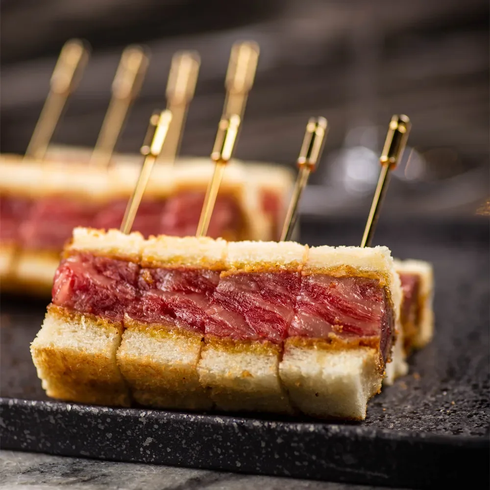 Close-up of beef tartare sandwich slices with toothpick skewers on a dark surface.