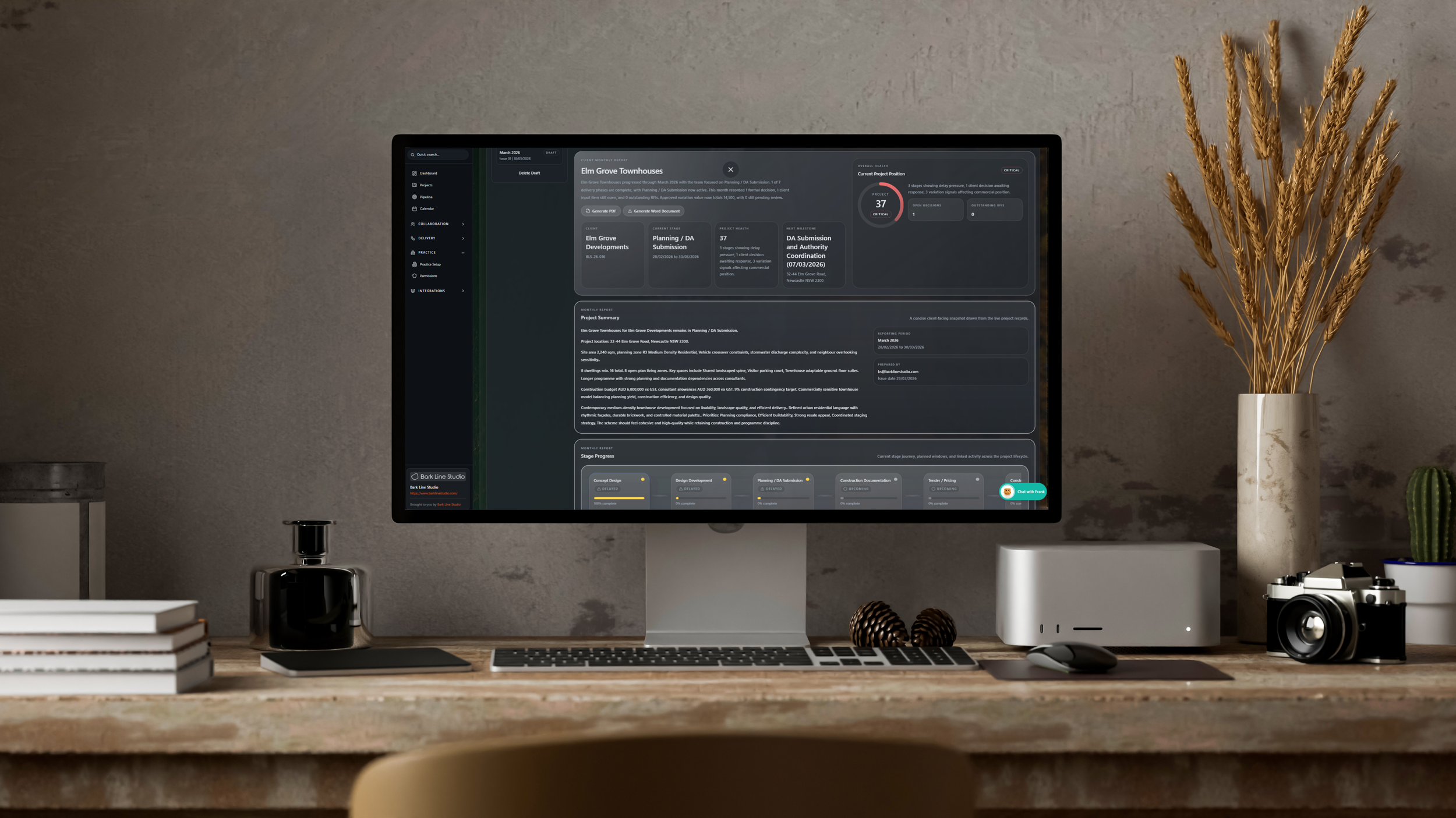 A computer monitor on a wooden desk displaying a project management dashboard with dark theme, alongside decorative items including a black bottle, a stack of white books, pine cones, a printer, a camera, potted cactus and dried plants in a tall beige vase, set against a textured wall.