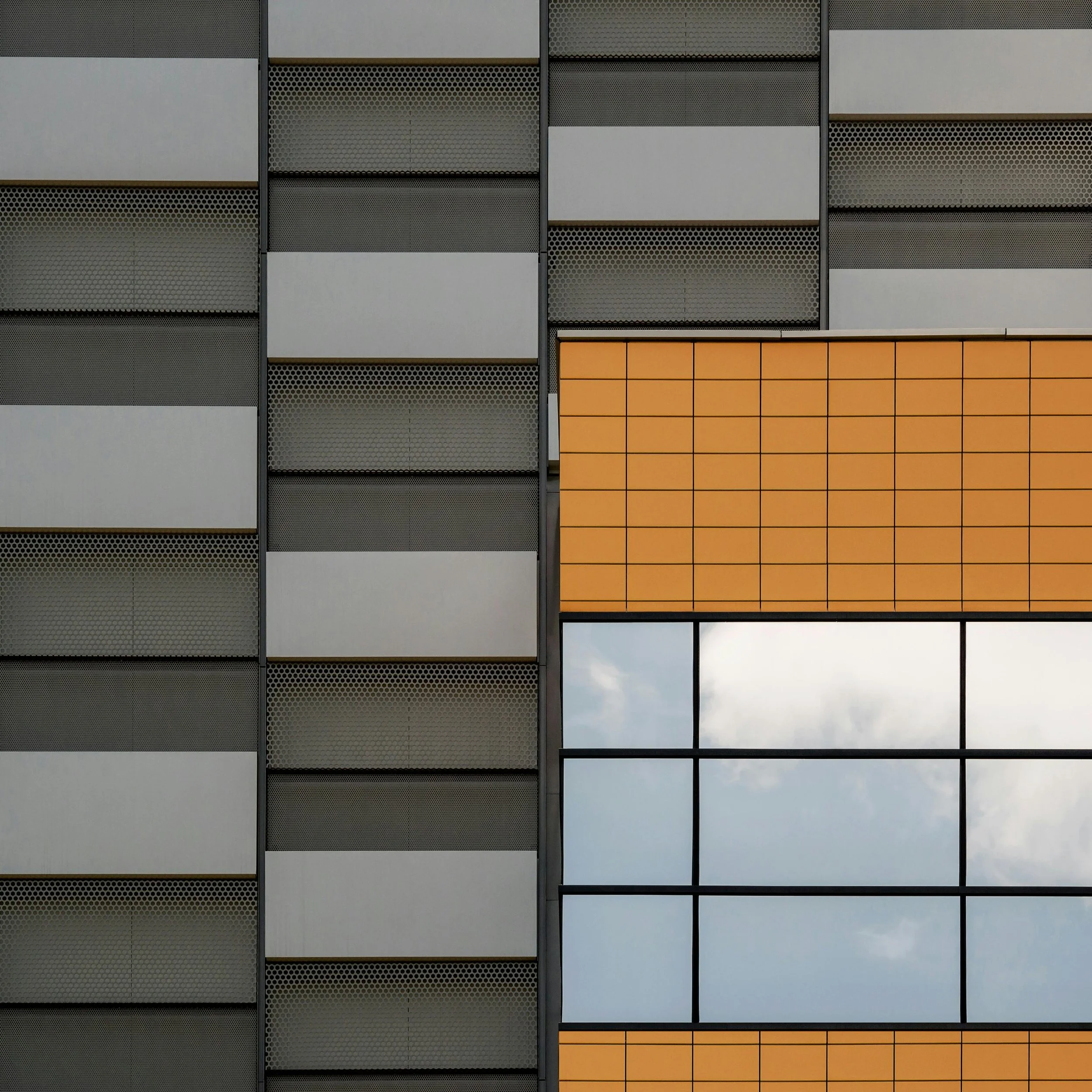 Close-up of a modern building facade with gray, black, and orange panels, featuring a grid of windows reflecting the sky.