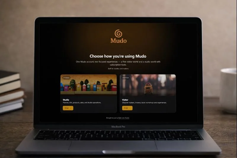 Laptop displaying the Mudo platform with options for Studio and Visitor modes on a desk with books to the left and a cup to the right.