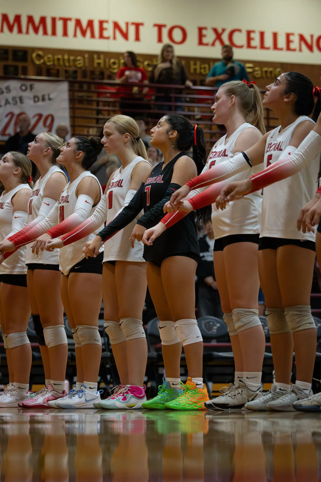 A volleyball team stands in a gymnasium during the national anthem, wearing white uniforms with one player in black, with red and white arm sleeves, and colorful athletic shoes.