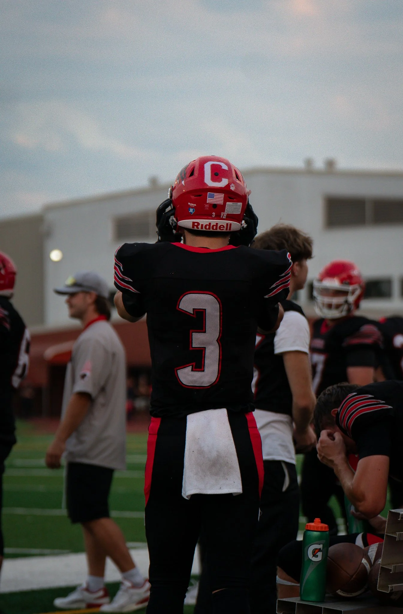 Football player wearing a red helmet with the letter 'C' on it and jersey number 3, standing on the sideline during a game, with teammates and a coach visible in the background.