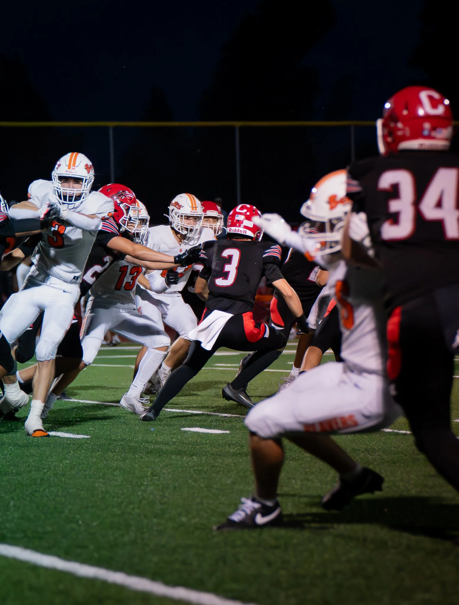 Youth football game at night with players on the field, some in white uniforms and others in black and red uniforms, engaging in a play.