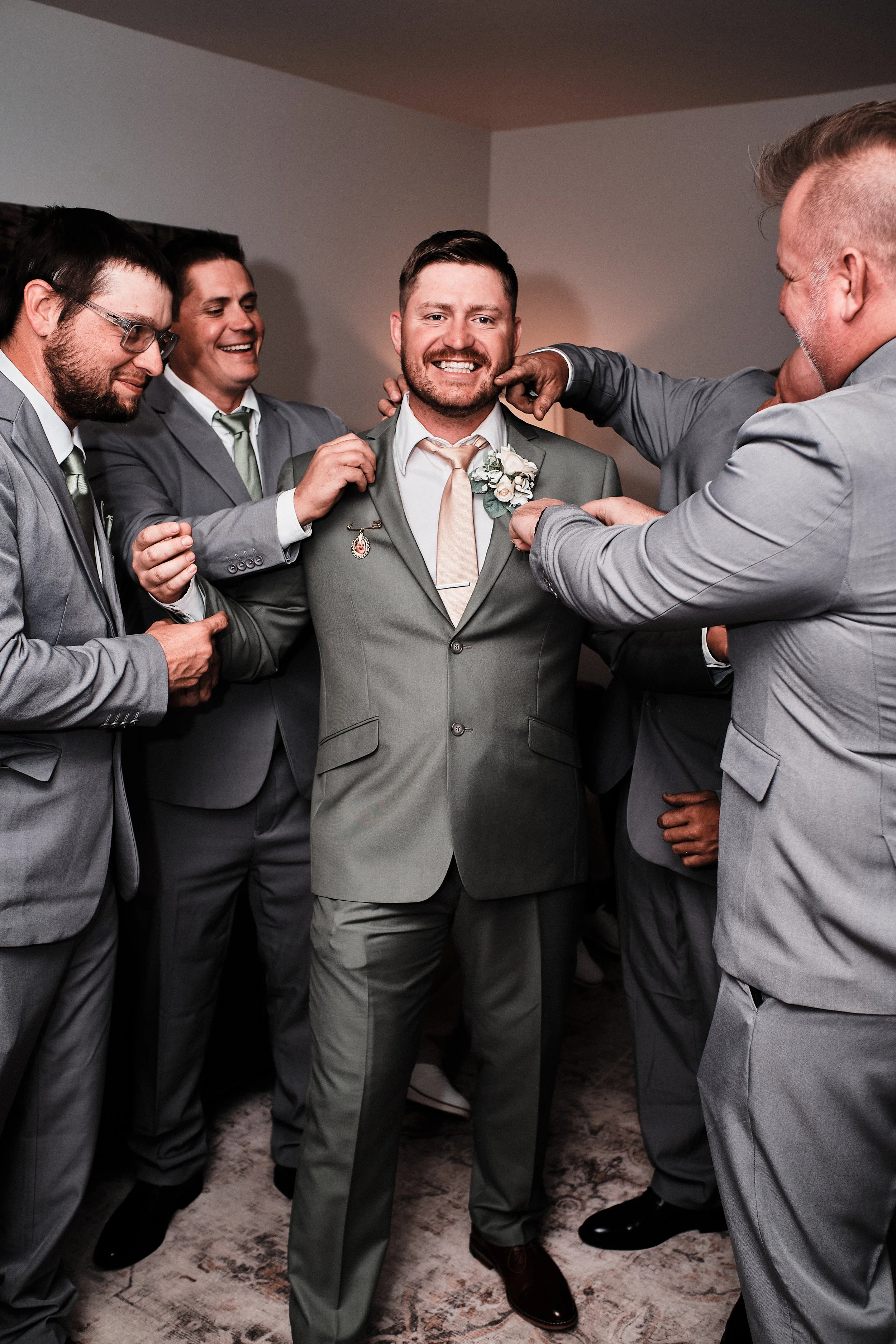 A group of men in gray suits and matching ties are smiling and having fun as they adjust a boutonniere on the smiling groom, who is at the center of the photo. The men are smiling and appear to be celebrating a wedding.