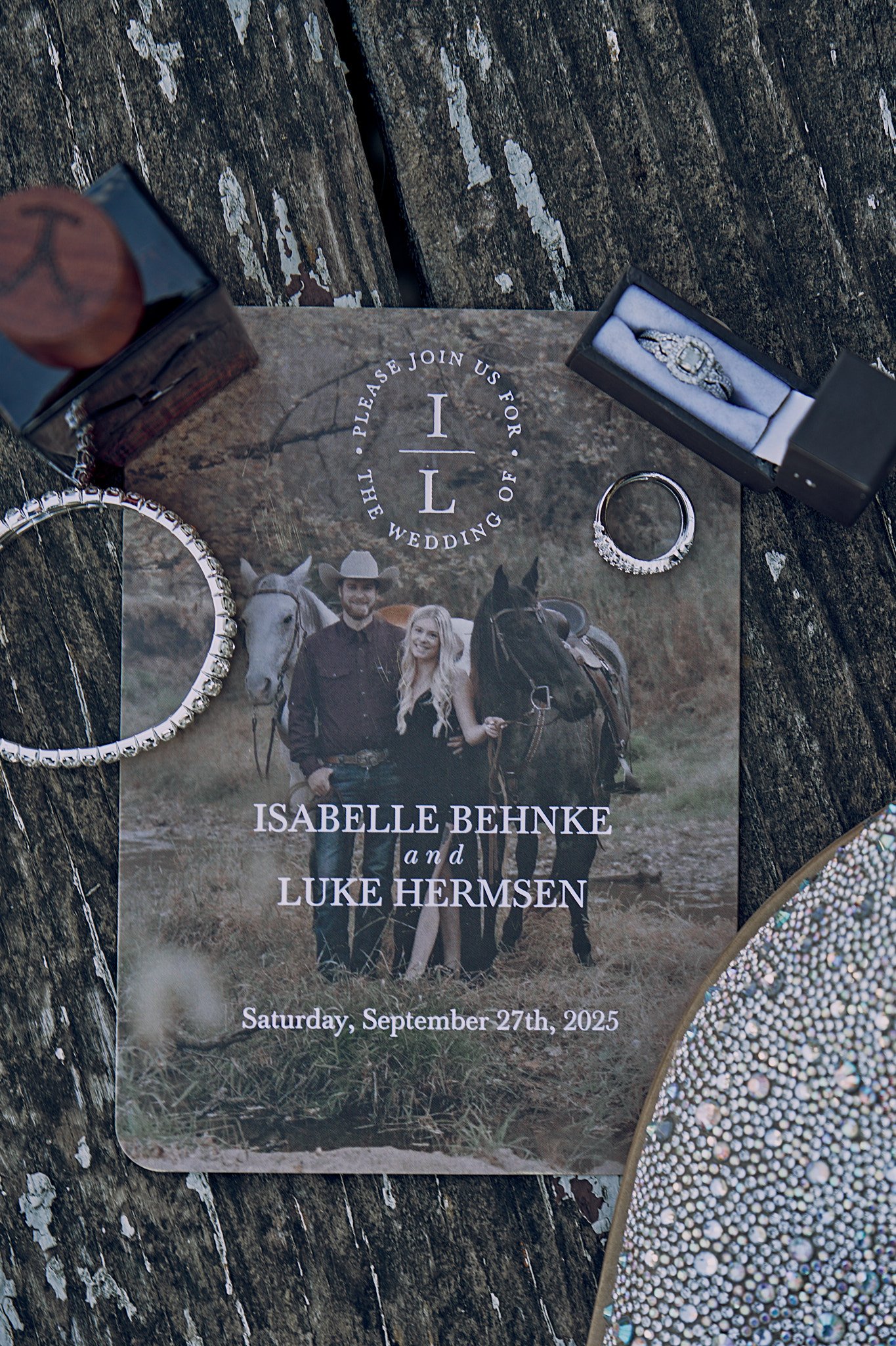 Wedding invitation with a photo of a couple with horses, surrounded by jewelry including a ring, a bracelet, and a watch, on a wooden surface.