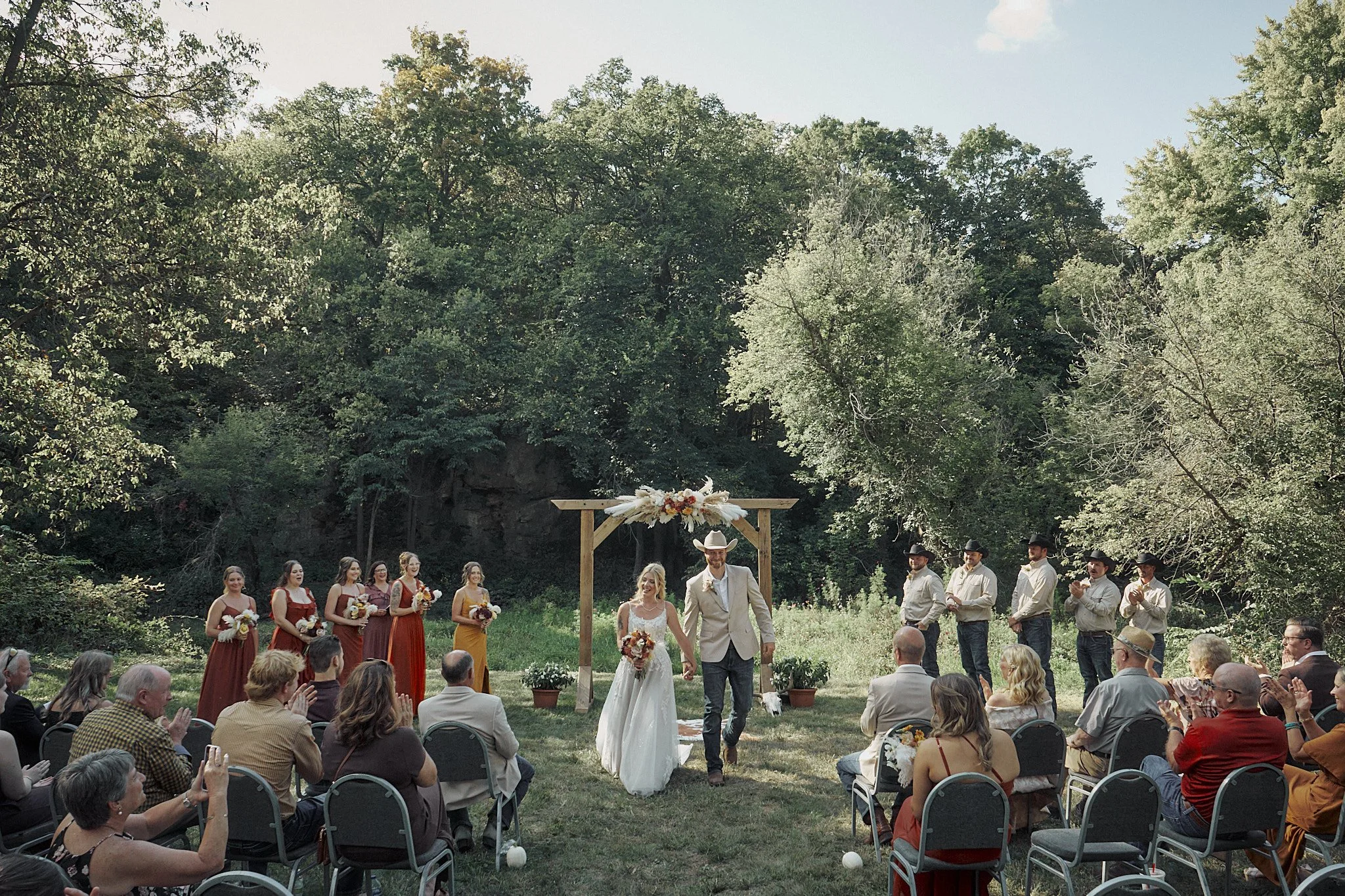 A couple walking down the aisle at an outdoor wedding ceremony in a forested area, with guests seated on either side and a decorated wooden arch in the background.