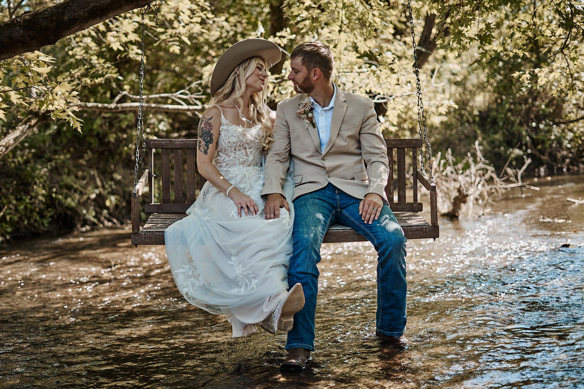 A couple sitting on a wooden swing over a creek, dressed in wedding attire, holding hands, and gazing at each other.