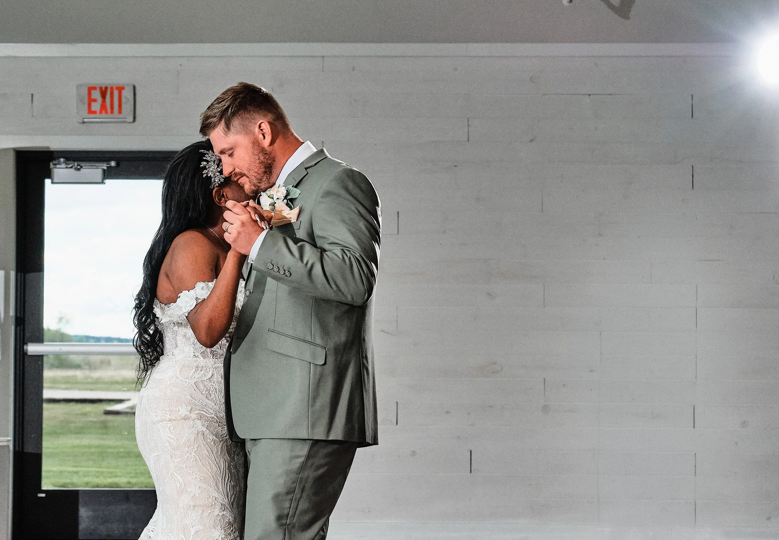 A bride and groom sharing a dance at their wedding, with the bride in a lace wedding dress and the groom in a gray suit, inside a modern building with a view of the outdoors.