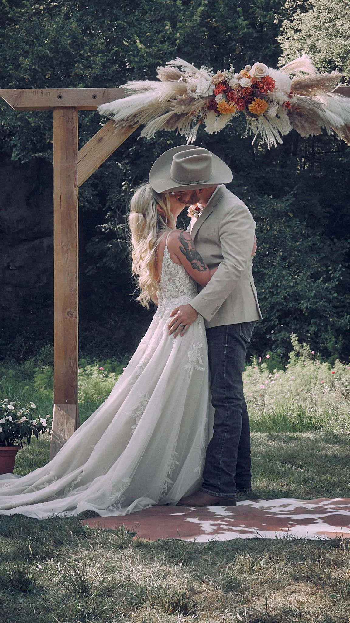 A couple kissing during their outdoor wedding ceremony under a wooden arch decorated with flowers and pampas grass.