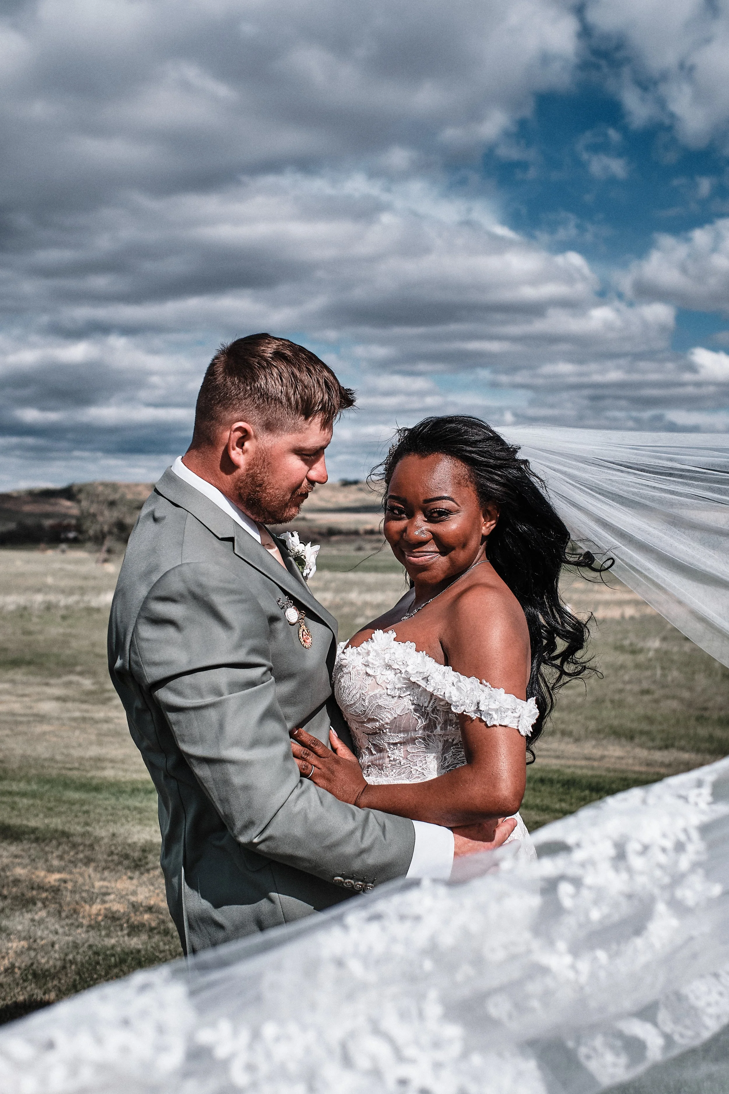A bride and groom standing close together outdoors on a cloudy day, with the groom in a gray suit and the bride in a white lace dress and veil, holding hands and smiling.