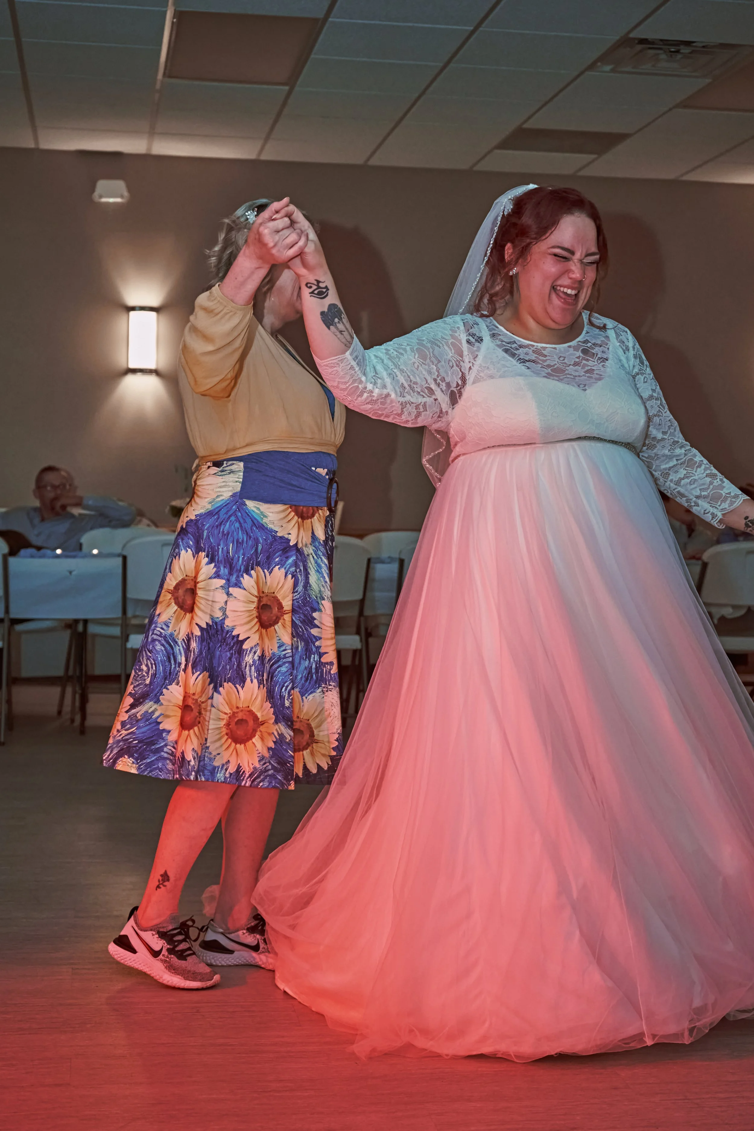 A bride in a pink and white wedding dress dancing with a guest in a flowering skirt and sneakers at a wedding reception.