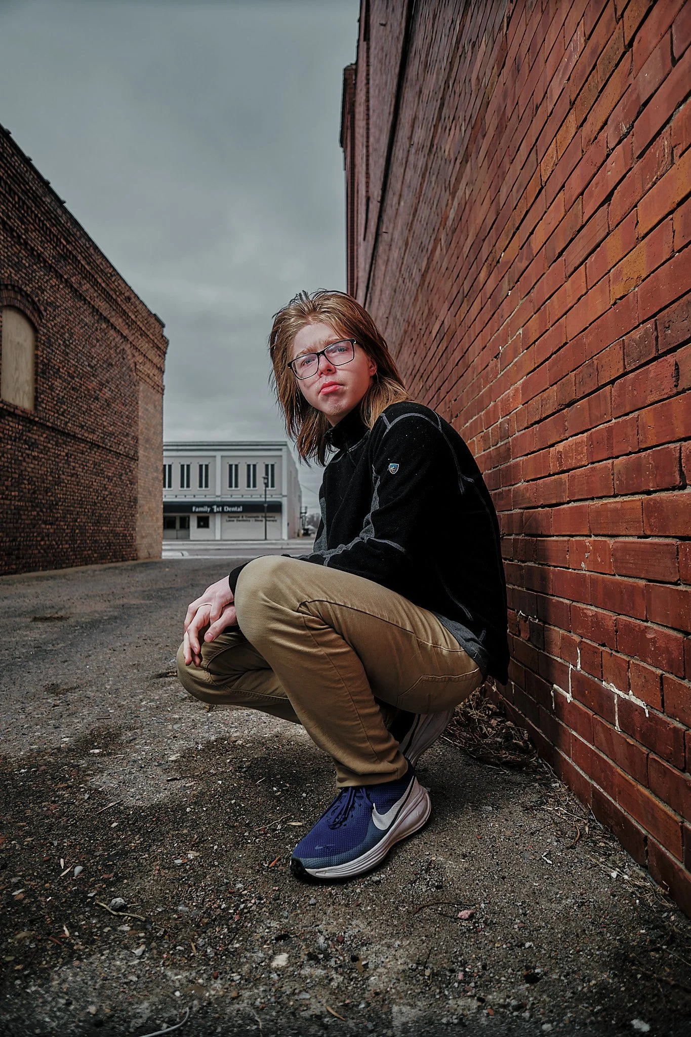 Young man crouching in an alley with brick walls on either side, wearing glasses, a black jacket, tan pants, and blue Nike sneakers, looking at the camera with a serious expression.