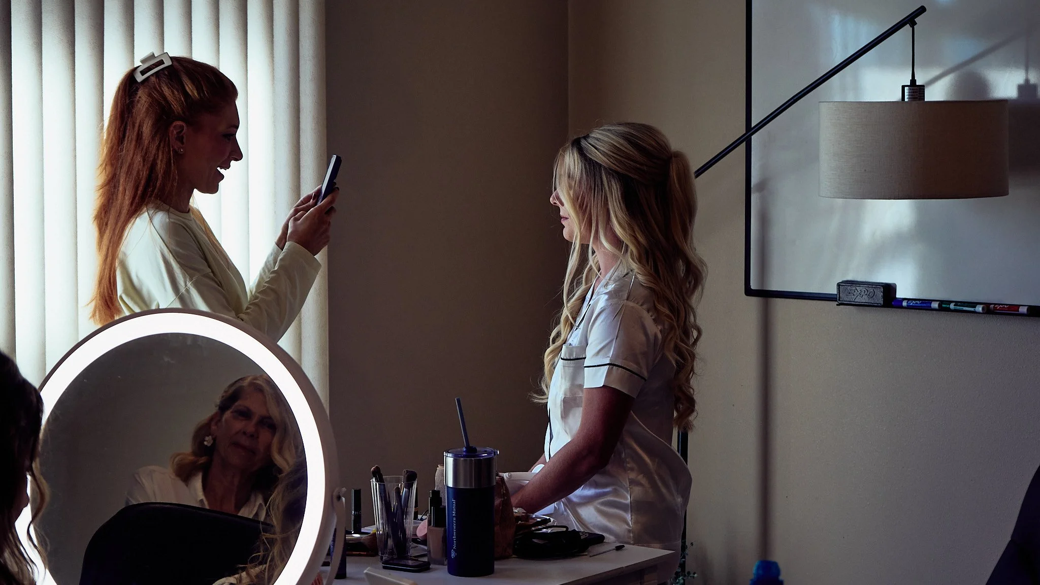 Two women in a makeup or skincare area, one with long blonde hair standing and looking at her phone, the other with long curly blonde hair sitting at a table with beauty products; a mirror with a built-in ring light reflects the seated woman.