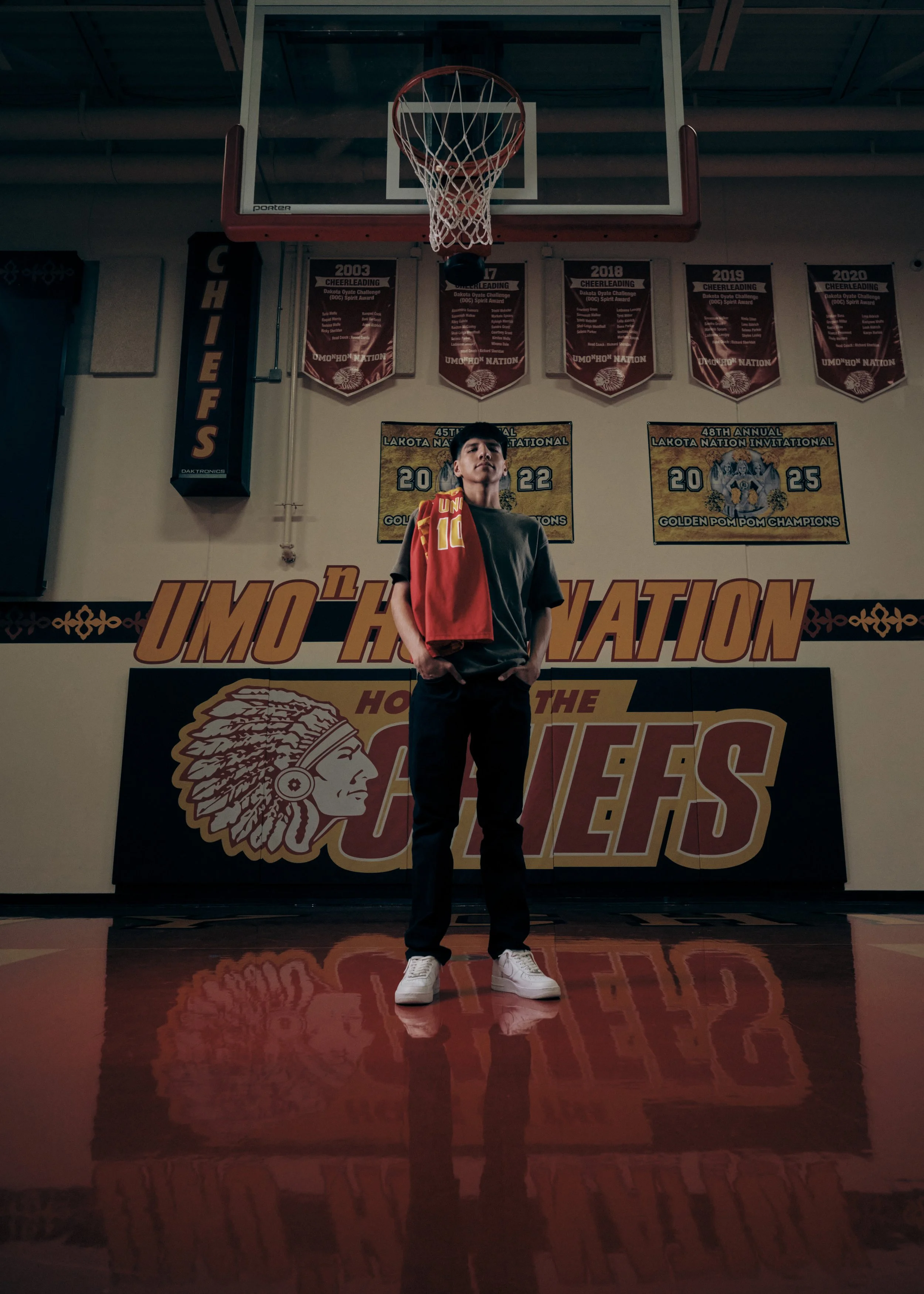 A young man standing in a gymnasium in front of a basketball hoop and championship banners. The background includes posters commemorating the 45th and 48th annual Lakota Nation Invitational, with a large logo of a Native American head. The man is wea