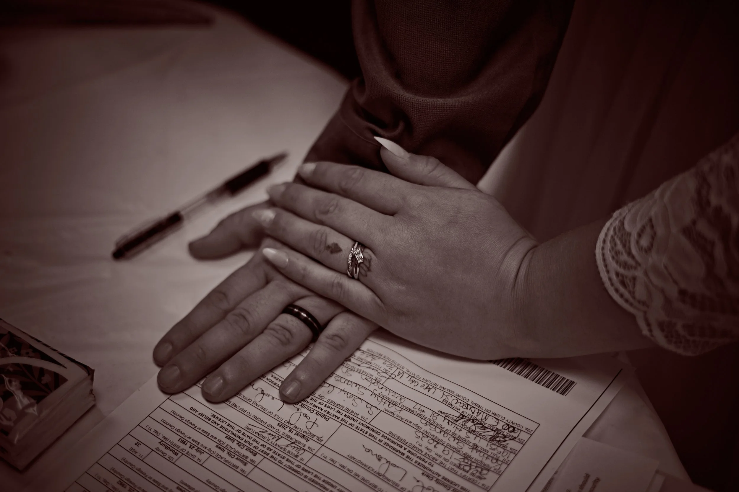 Two hands, one with a wedding ring and the other with an engagement ring, placed on top of a form on a table. Two pens are on the table, and the person is wearing a long-sleeved garment with lace detail.
