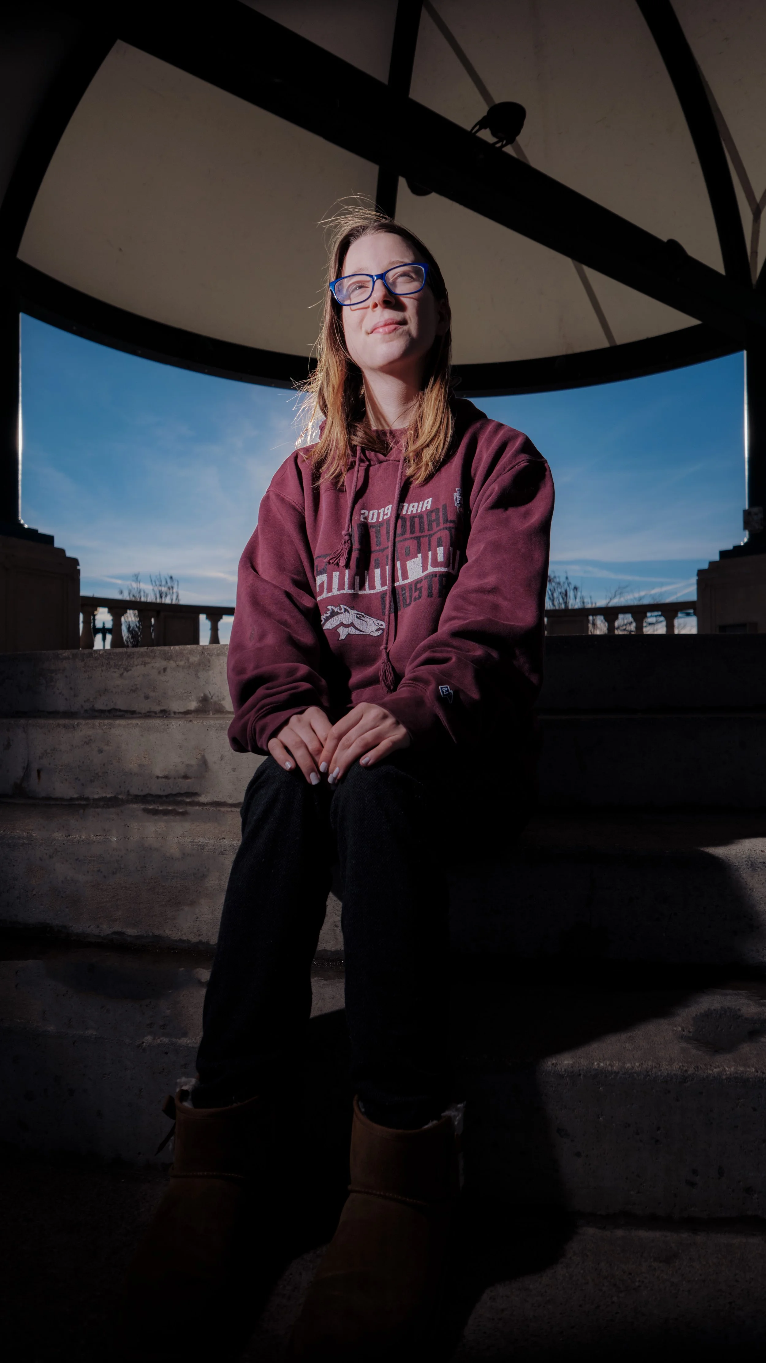 A young woman with glasses sitting on steps outdoors near a large clock structure, with a blue sky in the background.