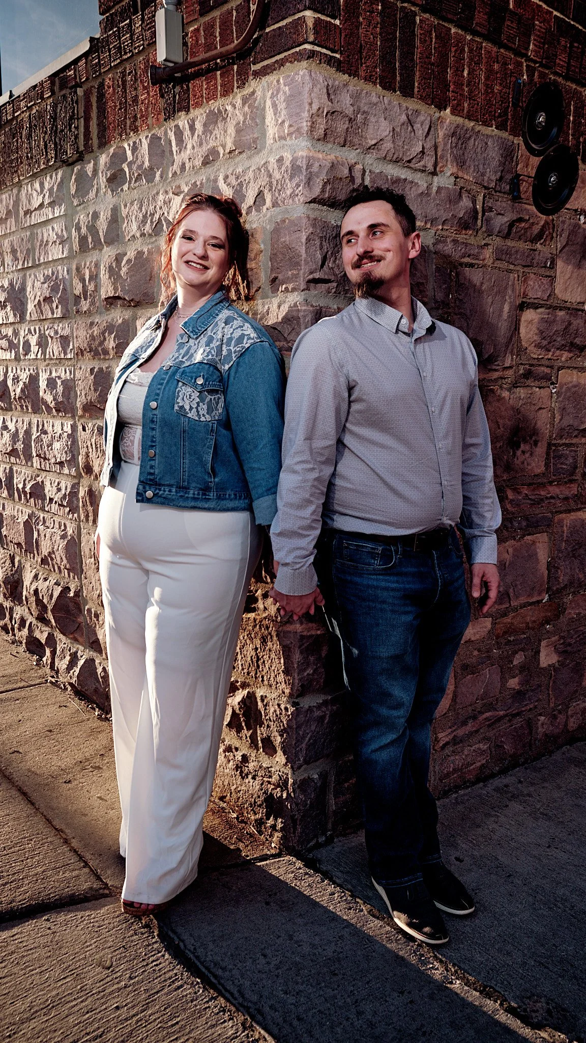 A couple holding hands, standing back to back against a brick wall, smiling and looking away from each other, outdoors during sunset with warm lighting.