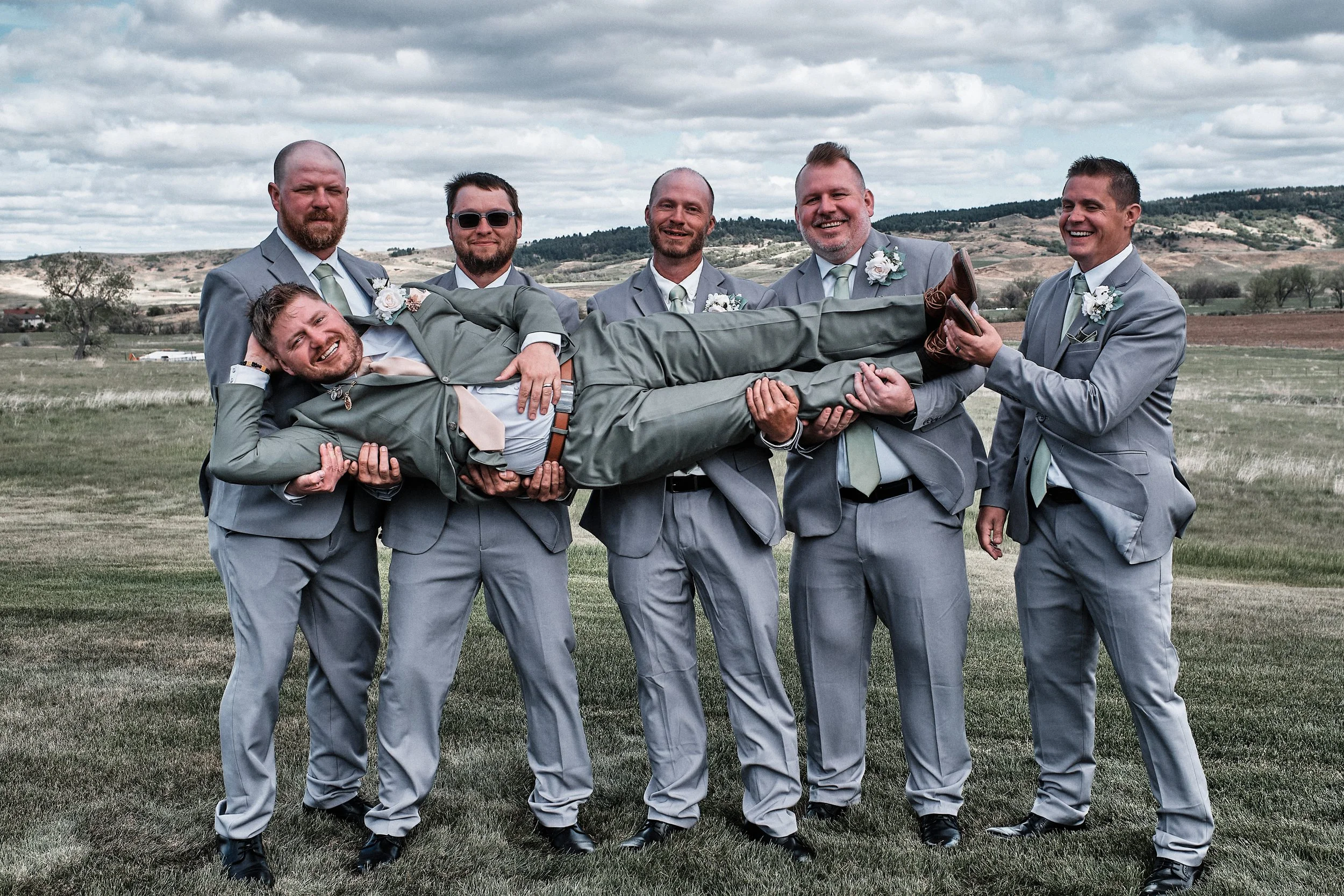 Seven men dressed in gray suits with light green ties and boutonnières, holding one man horizontally in a grassy field with hills and trees in the background, under a cloudy sky. All men are smiling or laughing.