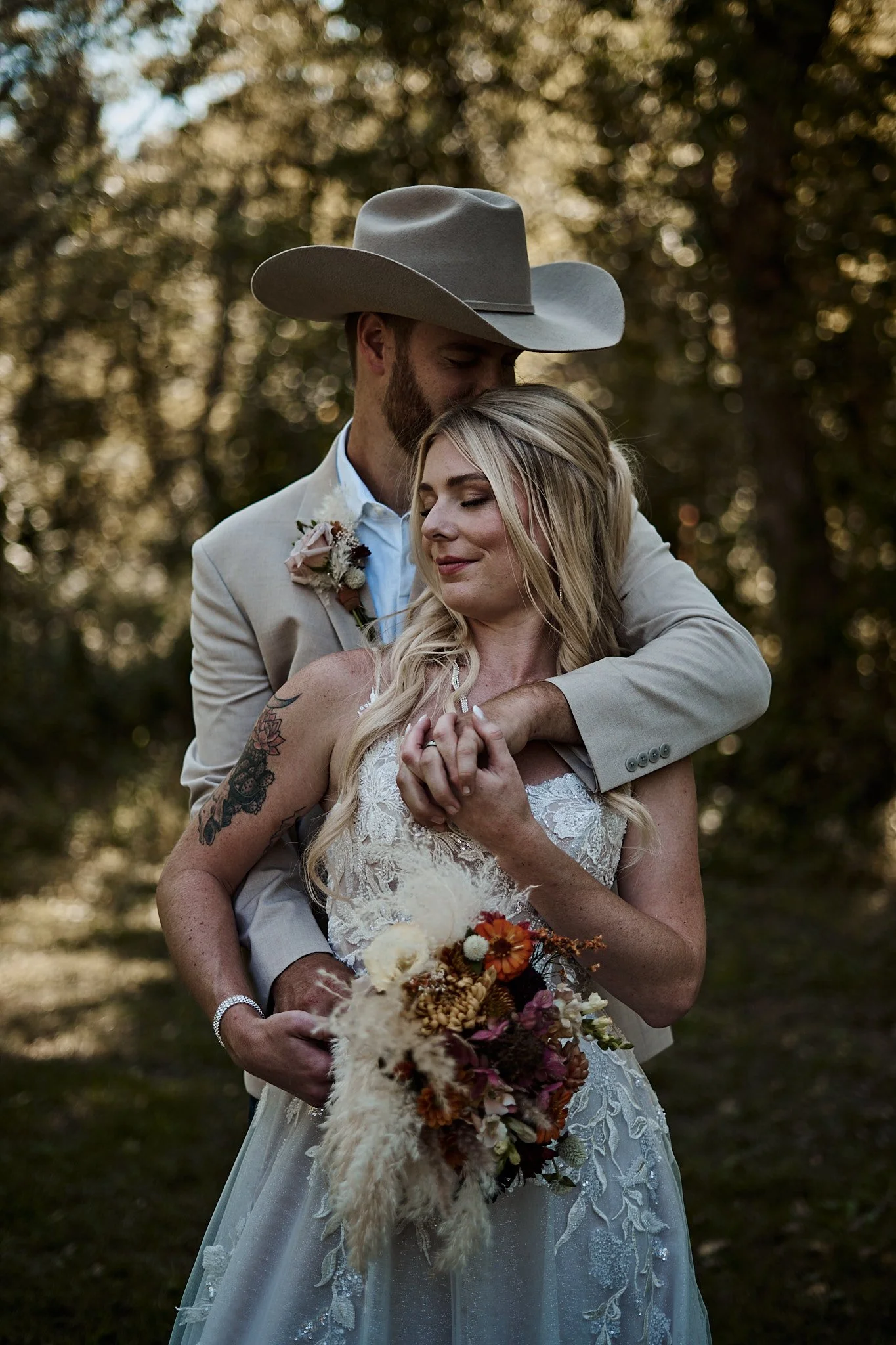 A couple in wedding attire sharing an intimate moment outdoors, with the groom embracing the bride from behind, in a wooded area during sunset.