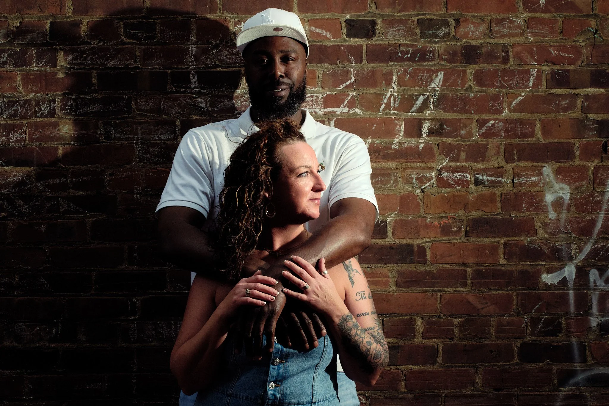 A man and a woman stand against a brick wall, with the man embracing the woman from behind.