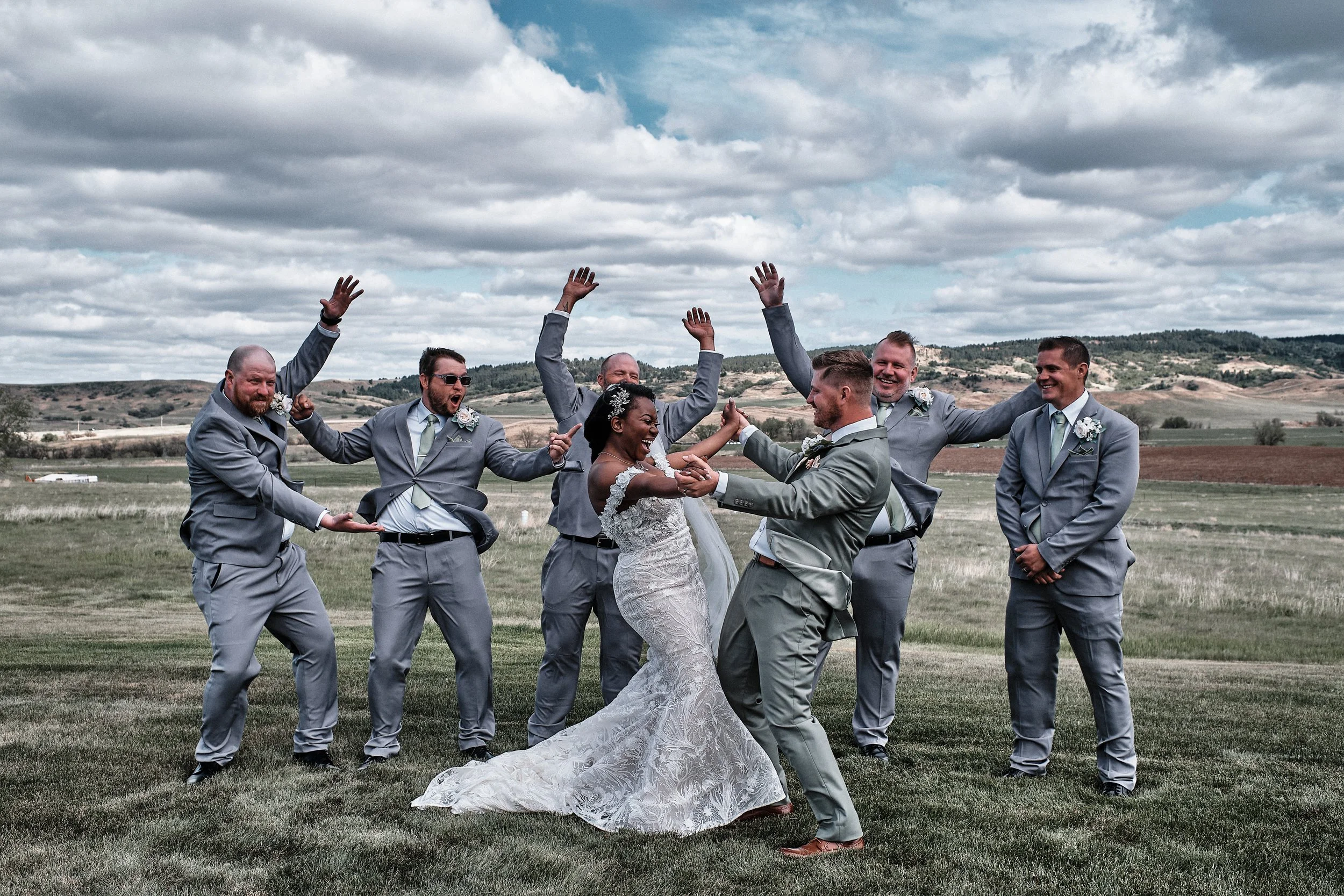 A bride in a white wedding dress dancing with a groom in a gray suit, surrounded by five groomsmen in matching gray suits, in an outdoor field under a cloudy sky.