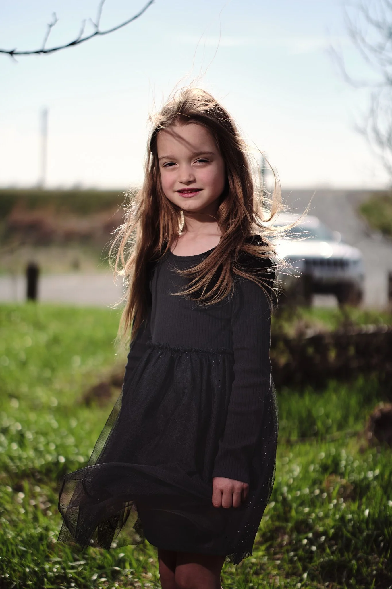 A young girl with long brown hair wearing a black dress standing outdoors on a grassy field with a blurry car and landscape in the background.