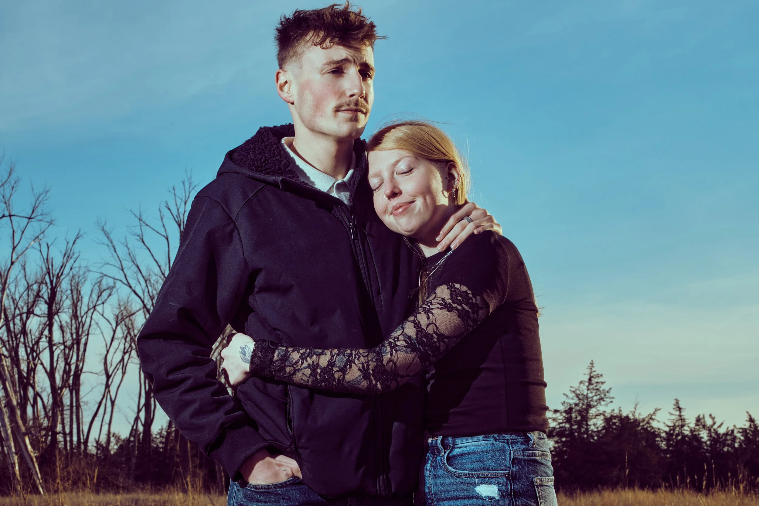 A couple stands outdoors, embracing lovingly. The man has short, dark hair, a mustache, and is wearing a black jacket. The woman has red hair, closed eyes, and is smiling softly as she leans into him. The background features a clear sky and leafless 