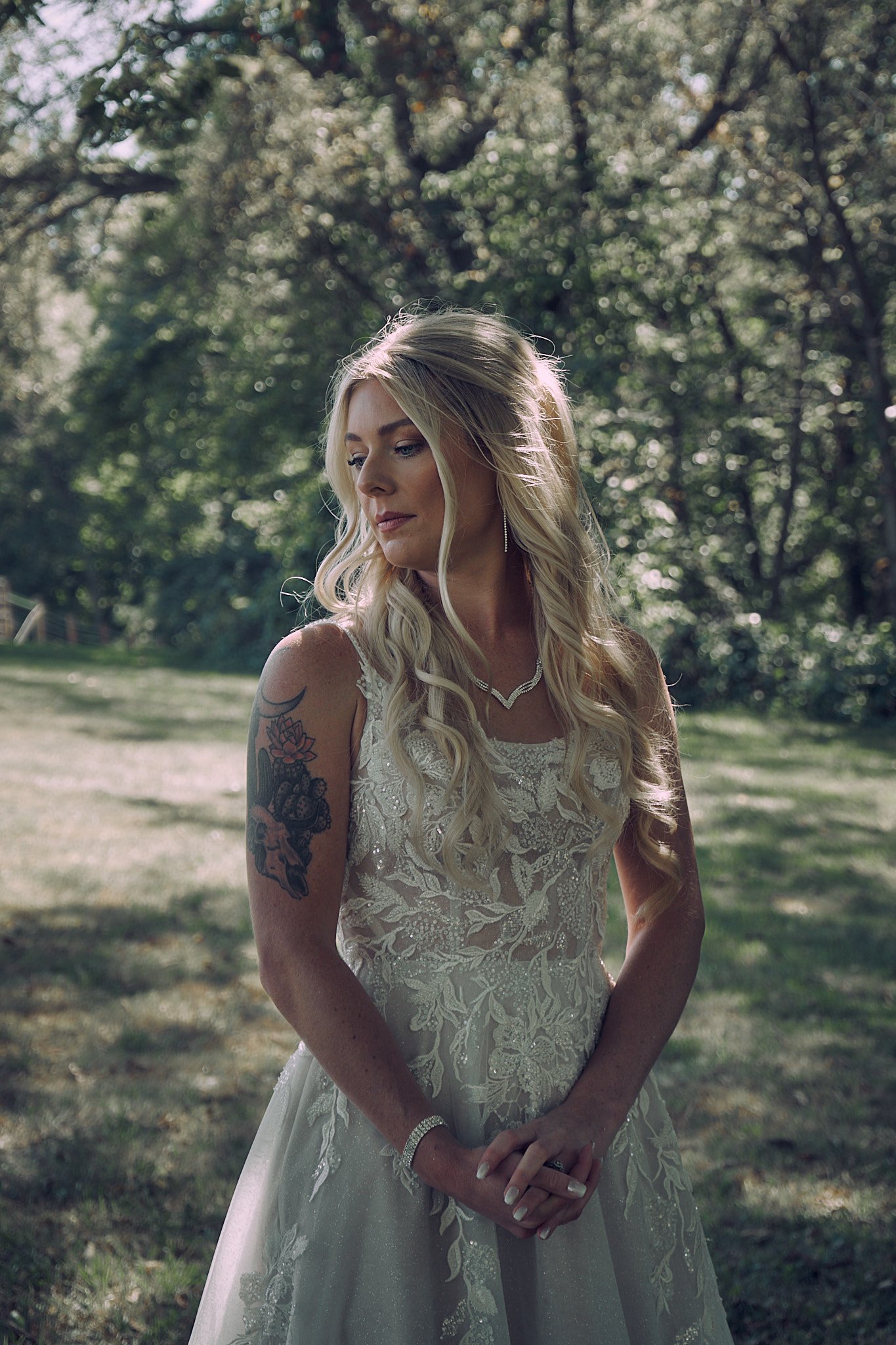 A bride in a lace wedding dress standing outdoors in a grassy area, with trees in the background. She has long blonde hair, tattoos on her left arm, and is wearing jewelry.