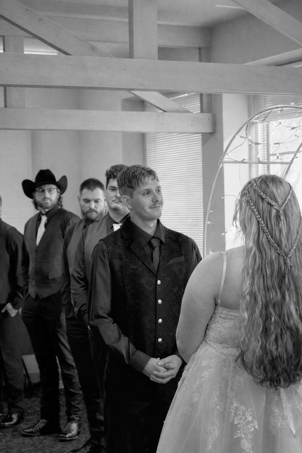 A wedding ceremony with a groom looking at the bride, who has long curly hair with braids, in a lace dress. Several men stand in a line behind the groom, some wearing suits and a cowboy hat, in an indoor space with natural light and decorative lights