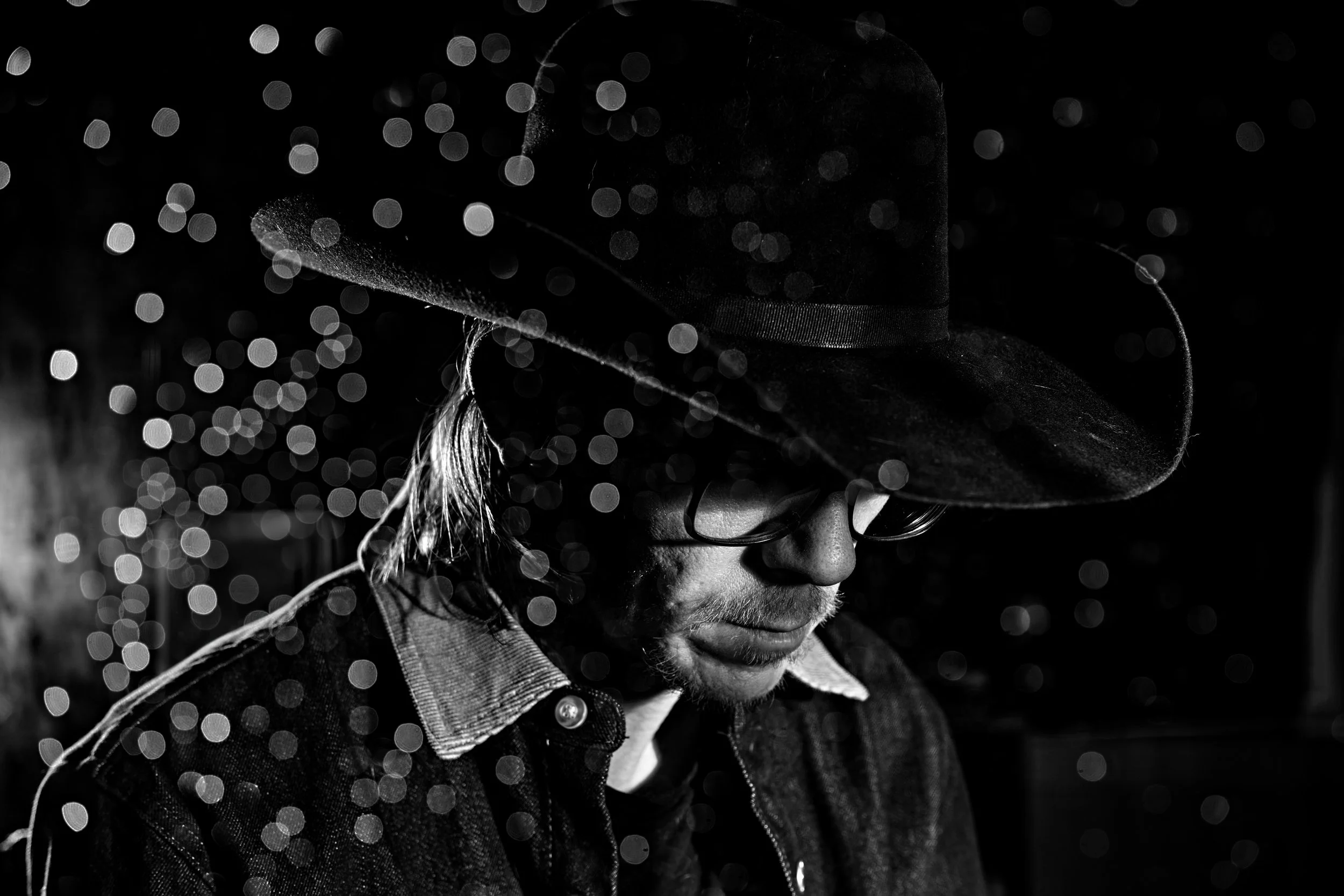 A black and white photo of a man wearing glasses and a wide-brimmed hat, with light reflections creating bokeh effects in the background.