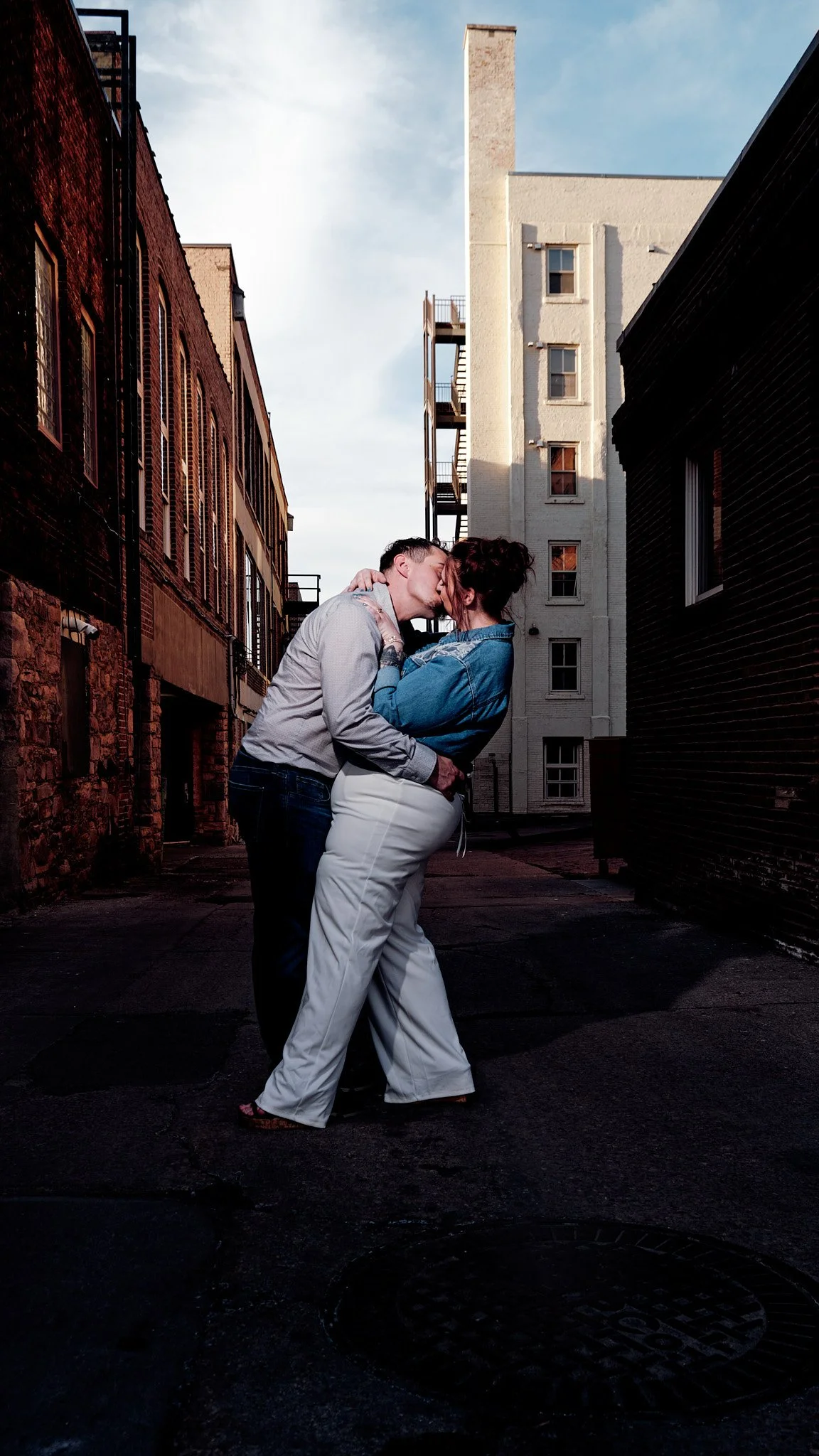 A couple kissing in an alleyway between tall brick buildings, with a partly cloudy sky overhead.