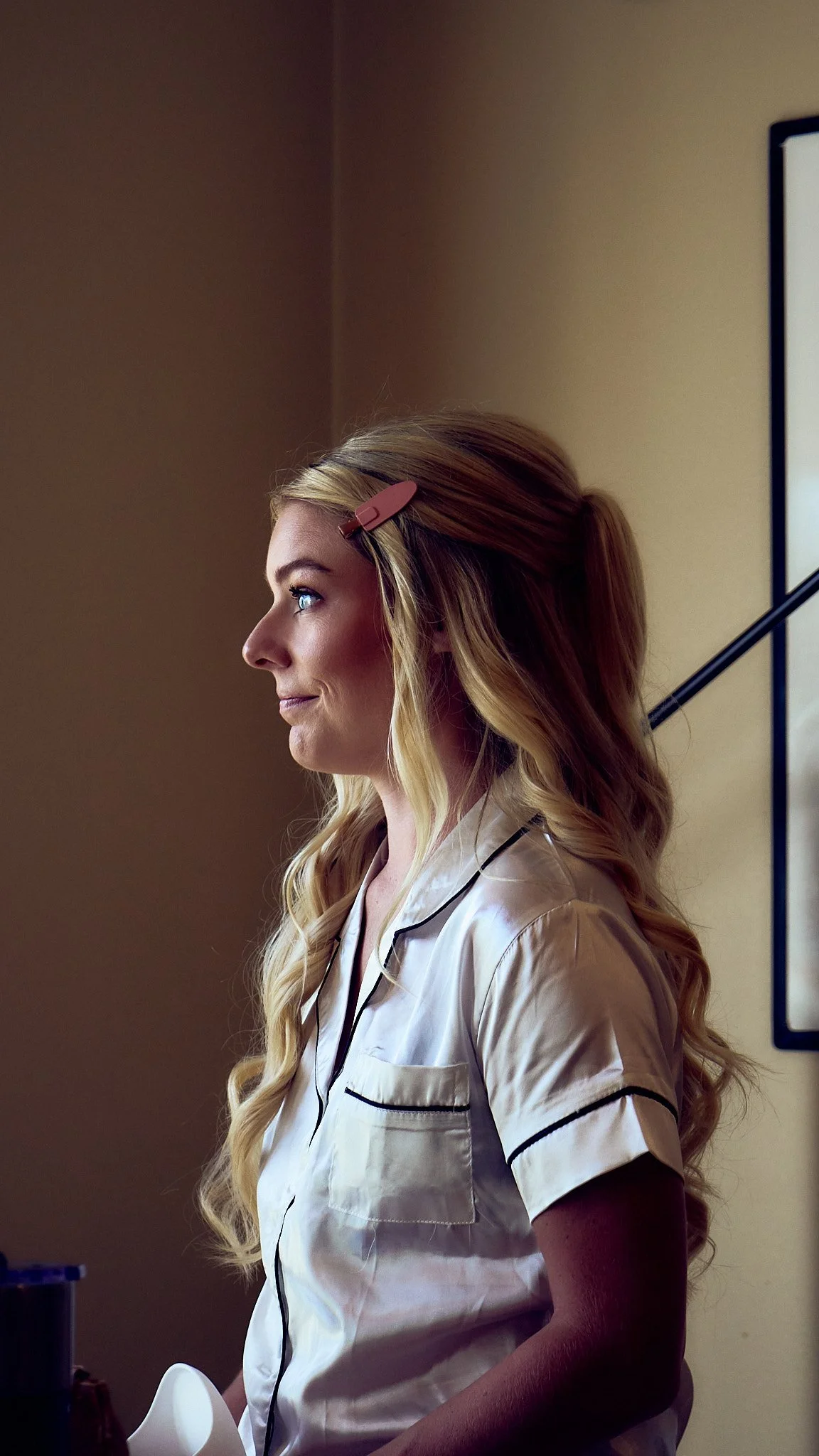 Side view of a woman with long blonde curly hair, wearing a pale-colored pajamas, sitting indoors.
