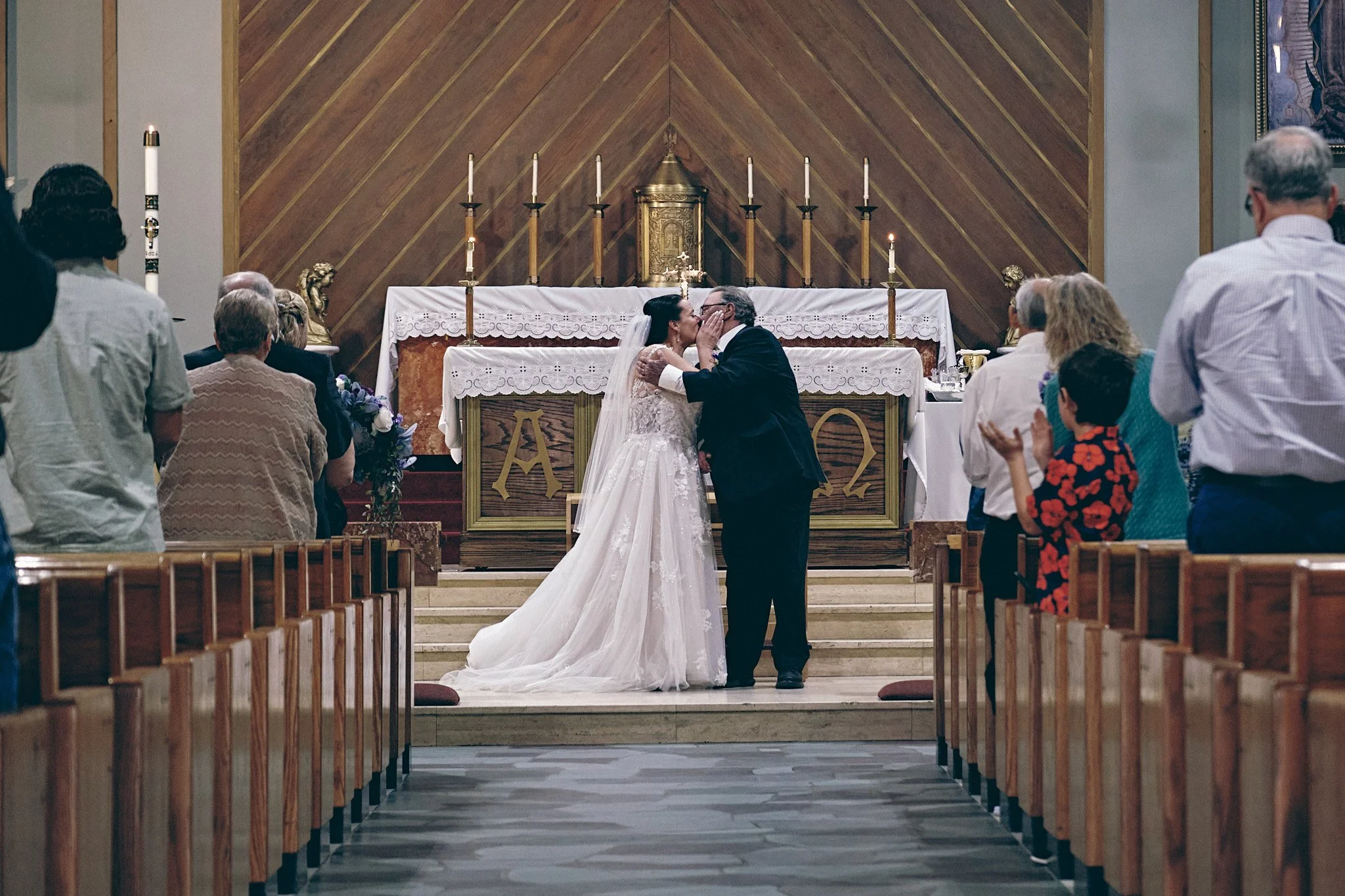 A bride and groom share a kiss at the altar during their wedding ceremony inside a church. The altar is decorated with candles and religious artifacts, with guests seated on wooden pews observing the moment.