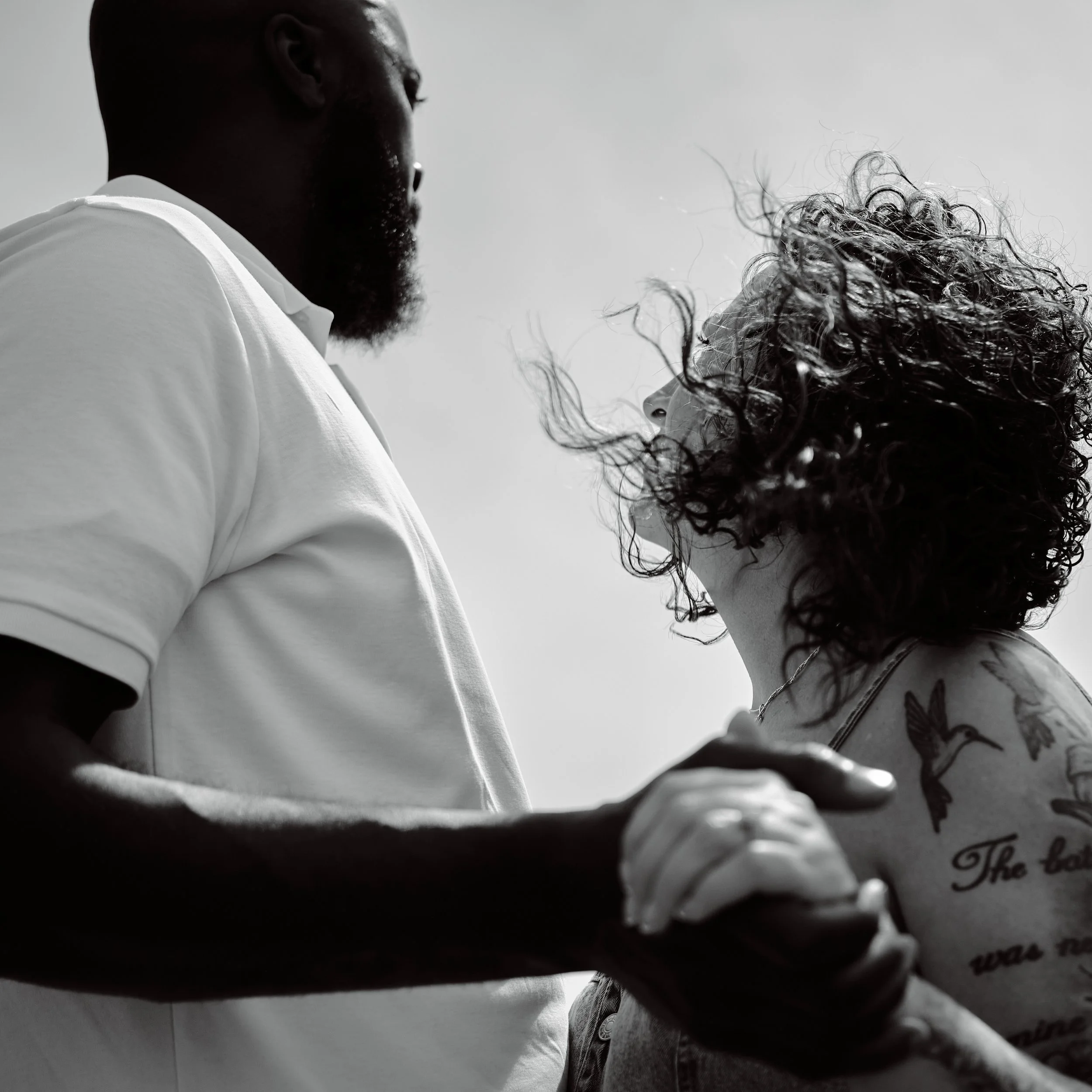 A black and white photo of a man and woman holding hands and looking into each other's eyes, with the man wearing a white shirt and the woman having curly hair and tattoos.