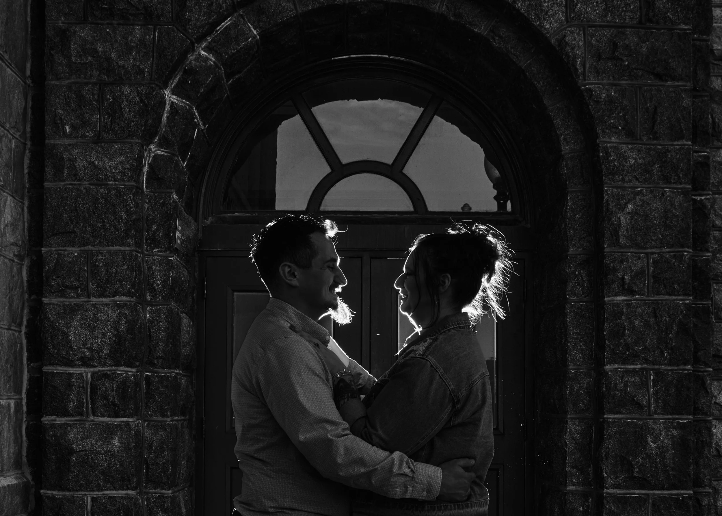 A black-and-white photo of a man and woman smiling at each other while embracing in front of a stone wall with an arched window behind them