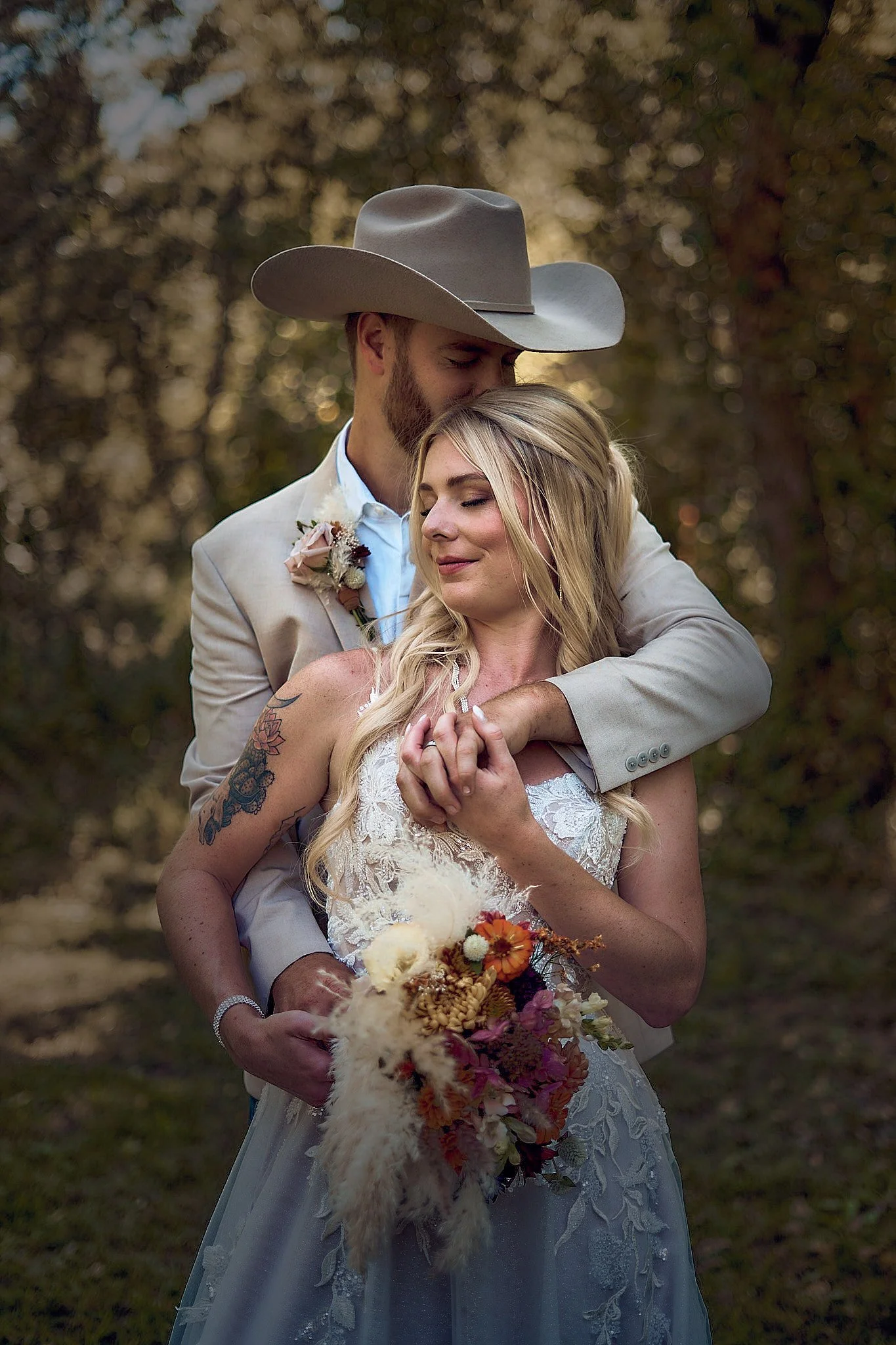 A newlywed couple hugging outdoors, with the groom wearing a cowboy hat, a beige suit, and a boutonniere, and the bride in a lace wedding dress holding a bouquet of flowers.