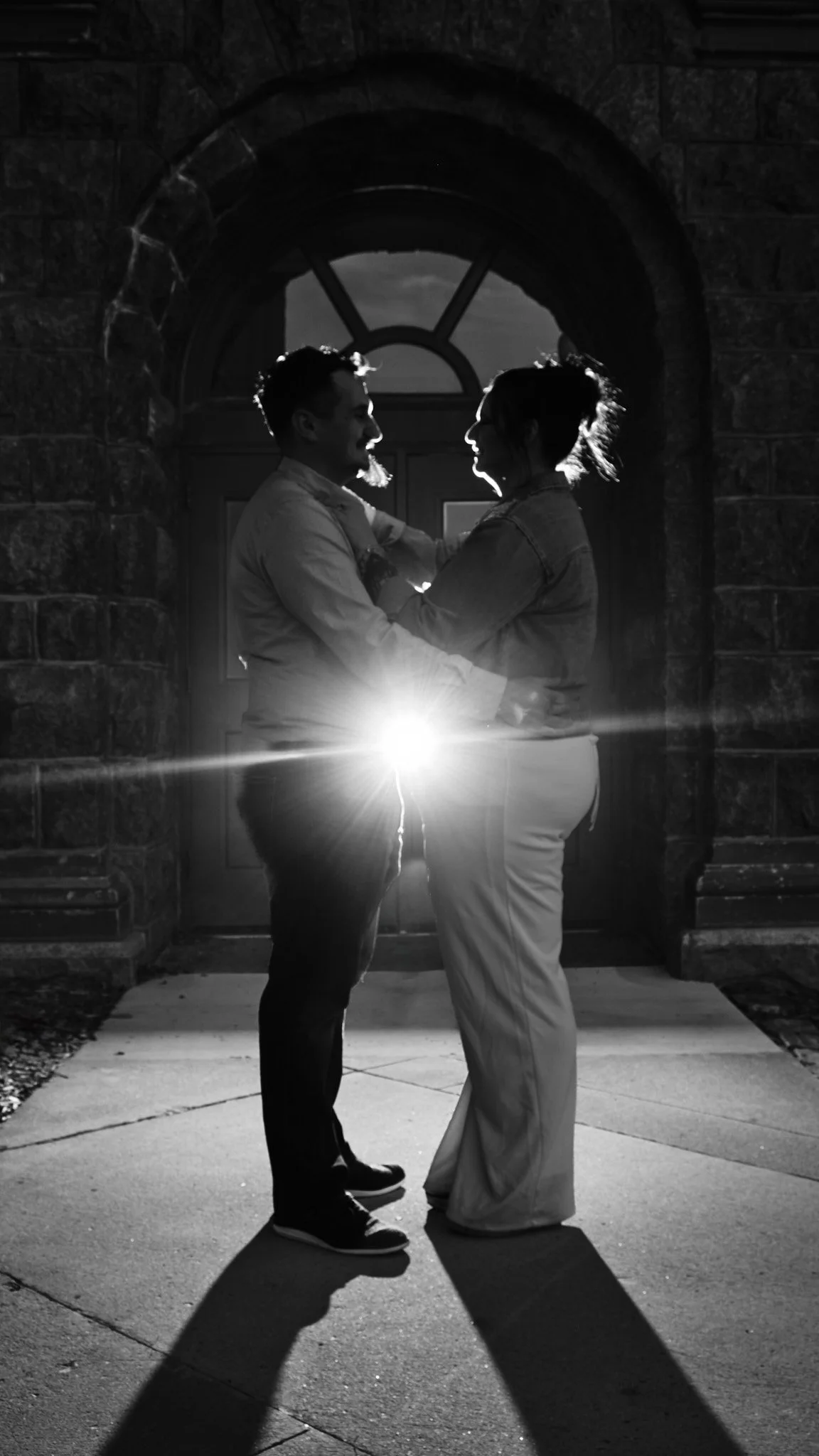 A black and white photo of a couple standing in front of a stone arch doorway at night, holding each other closely with the setting sun or a bright light behind them, casting their shadows on the ground.