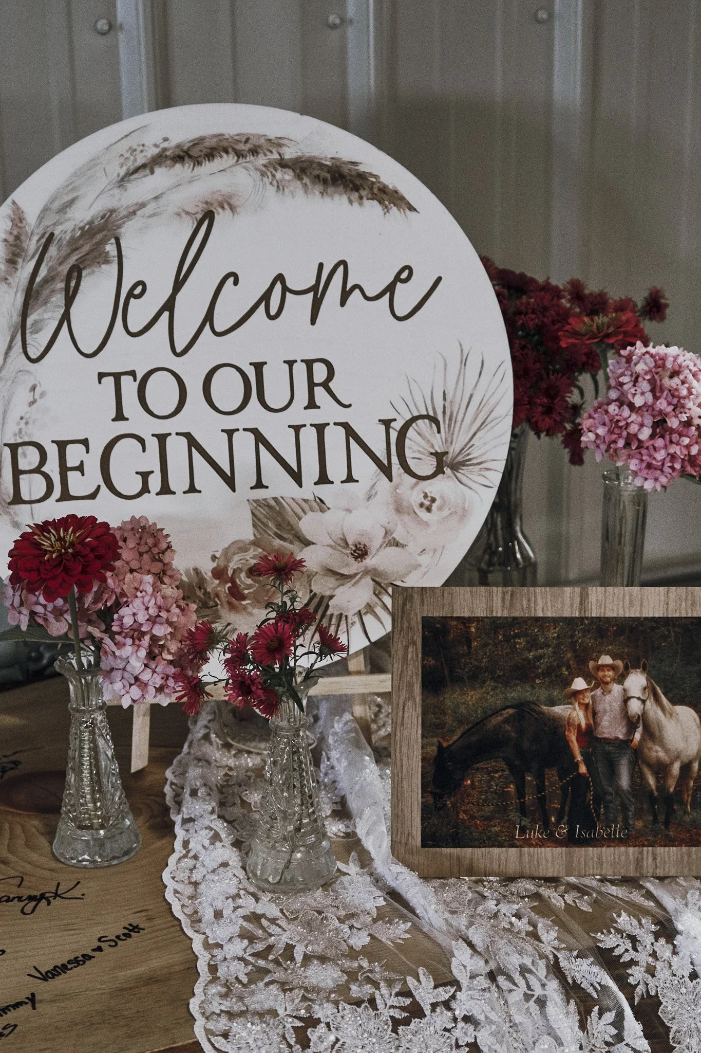 Decorative sign reading 'Welcome to our beginning' surrounded by pink and red flowers in glass vases and a framed picture of a couple with horses.
