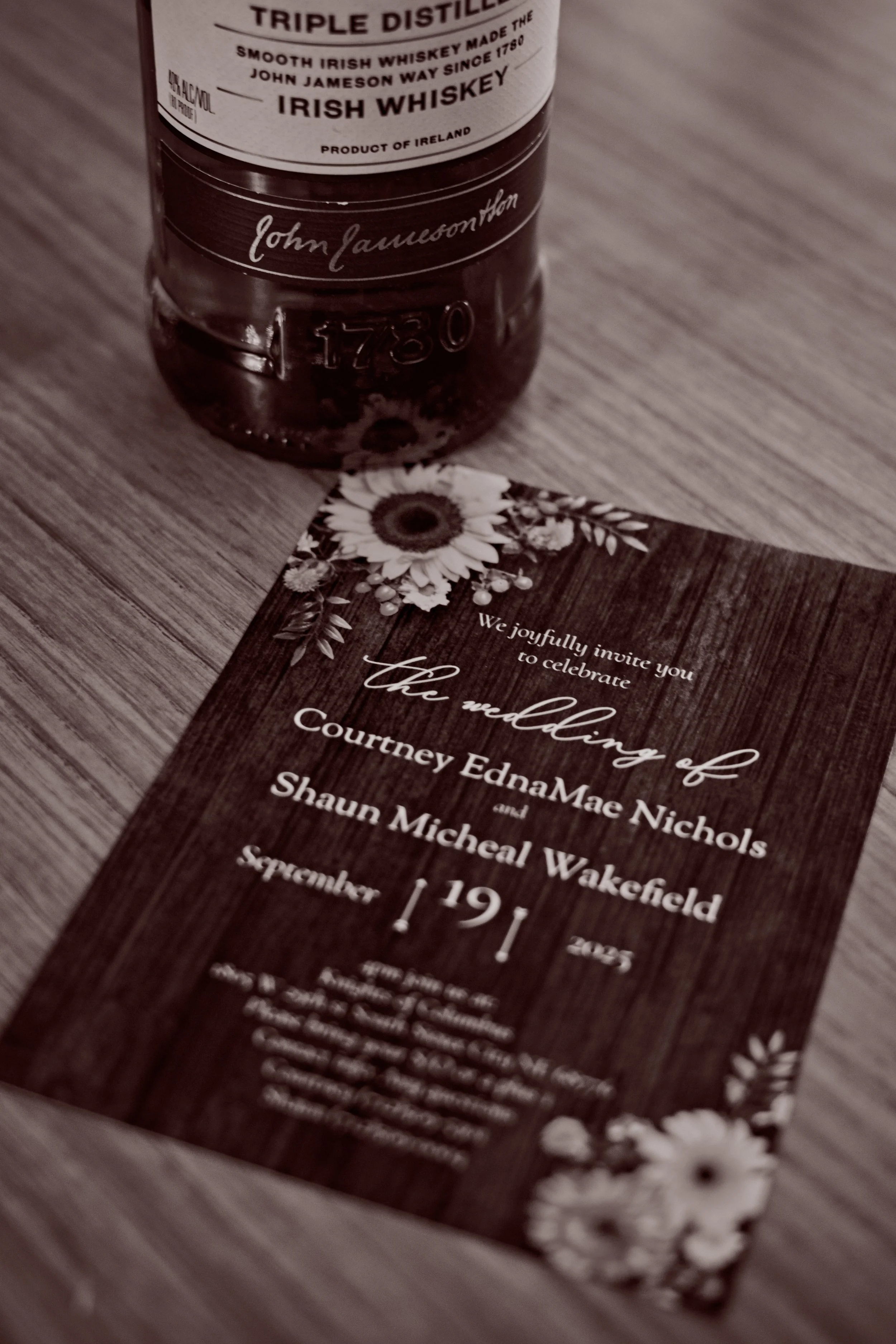 Wedding invitation card with floral decorations and an Irish whiskey bottle placed above it on a wooden surface.