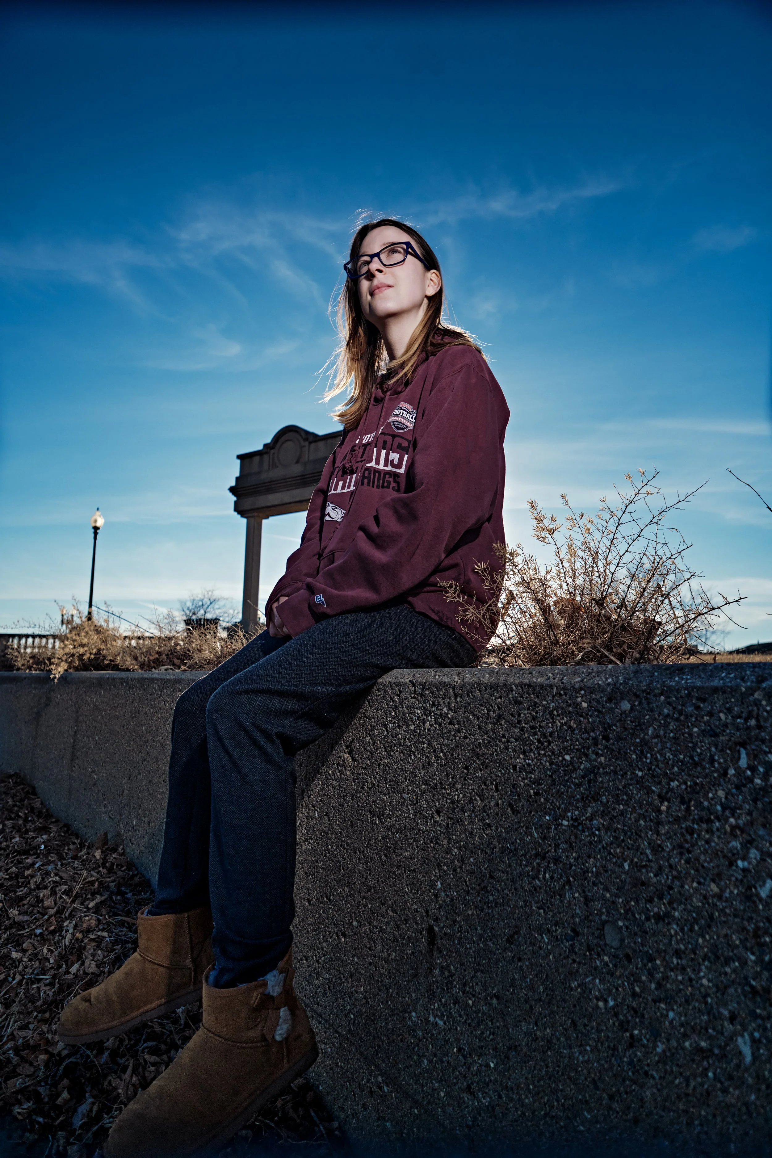 Young woman sitting on a concrete ledge outdoors, wearing glasses, a maroon jacket, dark pants, and brown boots, with a clear blue sky and some shrubbery behind her.