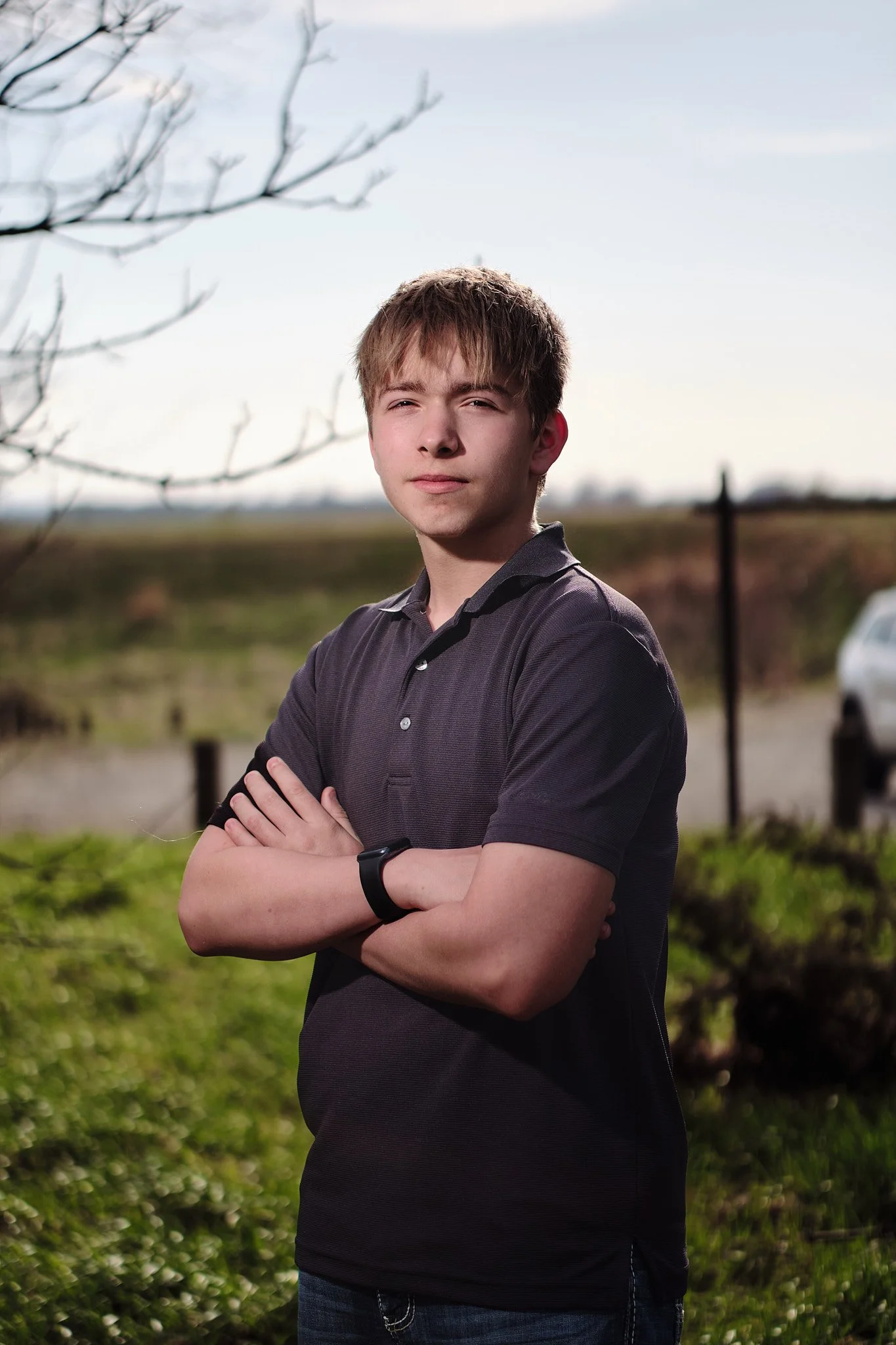 A young man with short light brown hair, wearing a dark gray polo shirt and a fitness tracker, stands outdoors with arms crossed, looking at the camera with a serious expression. The background includes a grassy area, a blurred car, and a partly clou