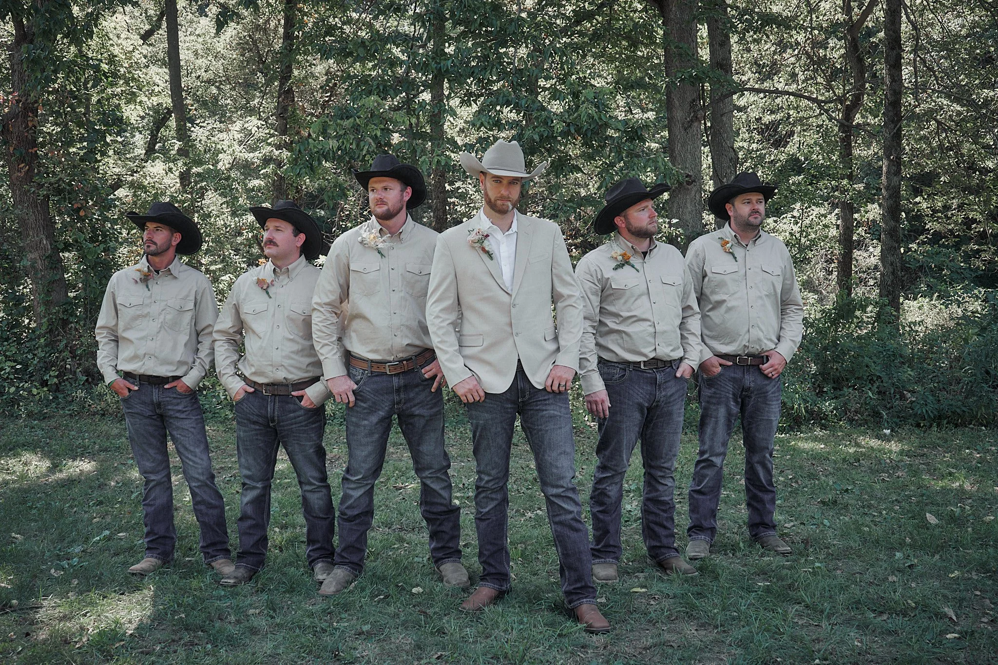 Group of six men standing outdoors in a row, dressed in casual western-style clothing, with five wearing black hats and one wearing a light gray hat, in a wooded area with trees and greenery in the background.