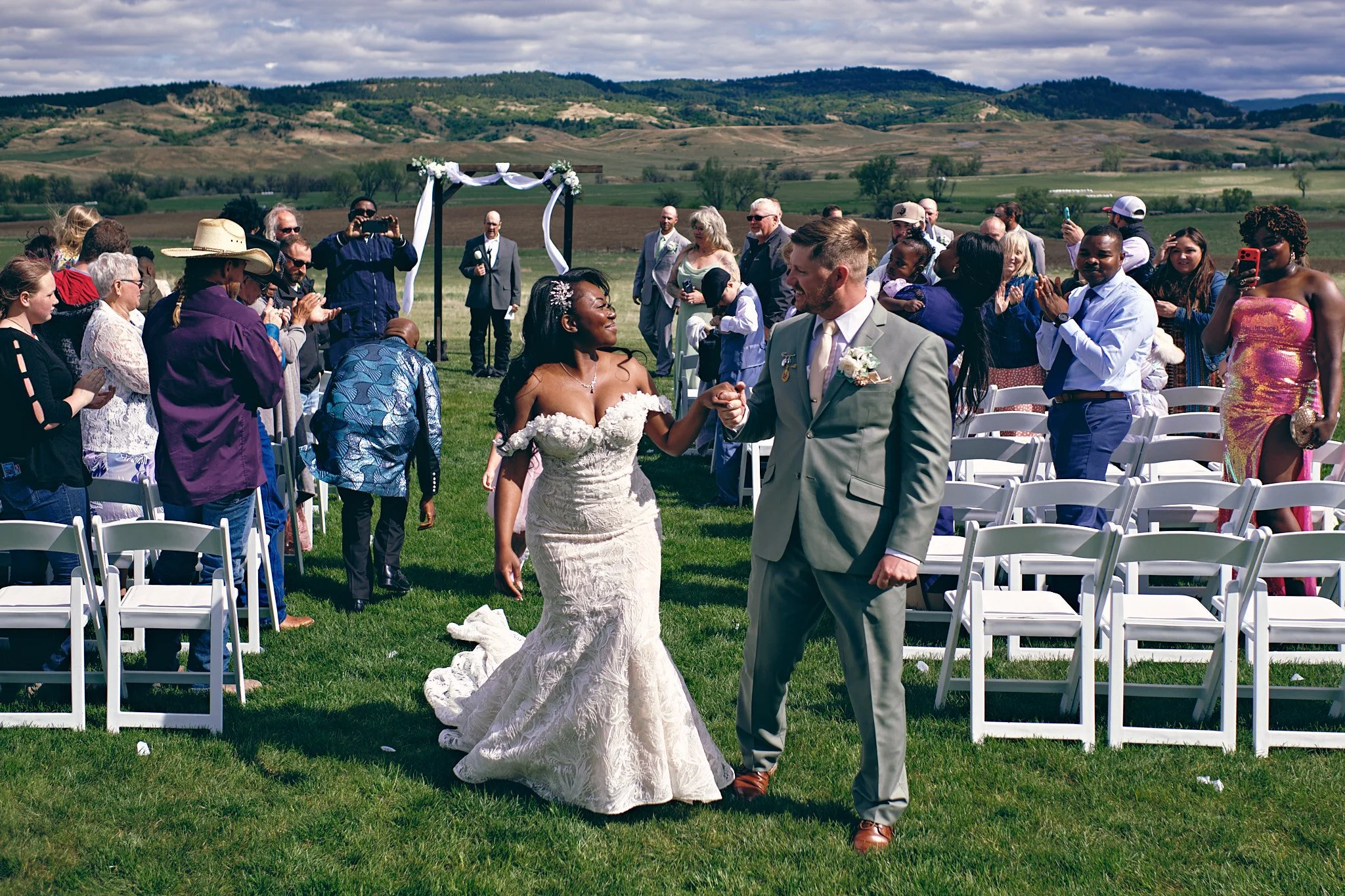 A bride and groom holding hands and walking down the aisle at an outdoor wedding ceremony with guests standing and clapping around them, set against a backdrop of rolling hills and a partly cloudy sky.